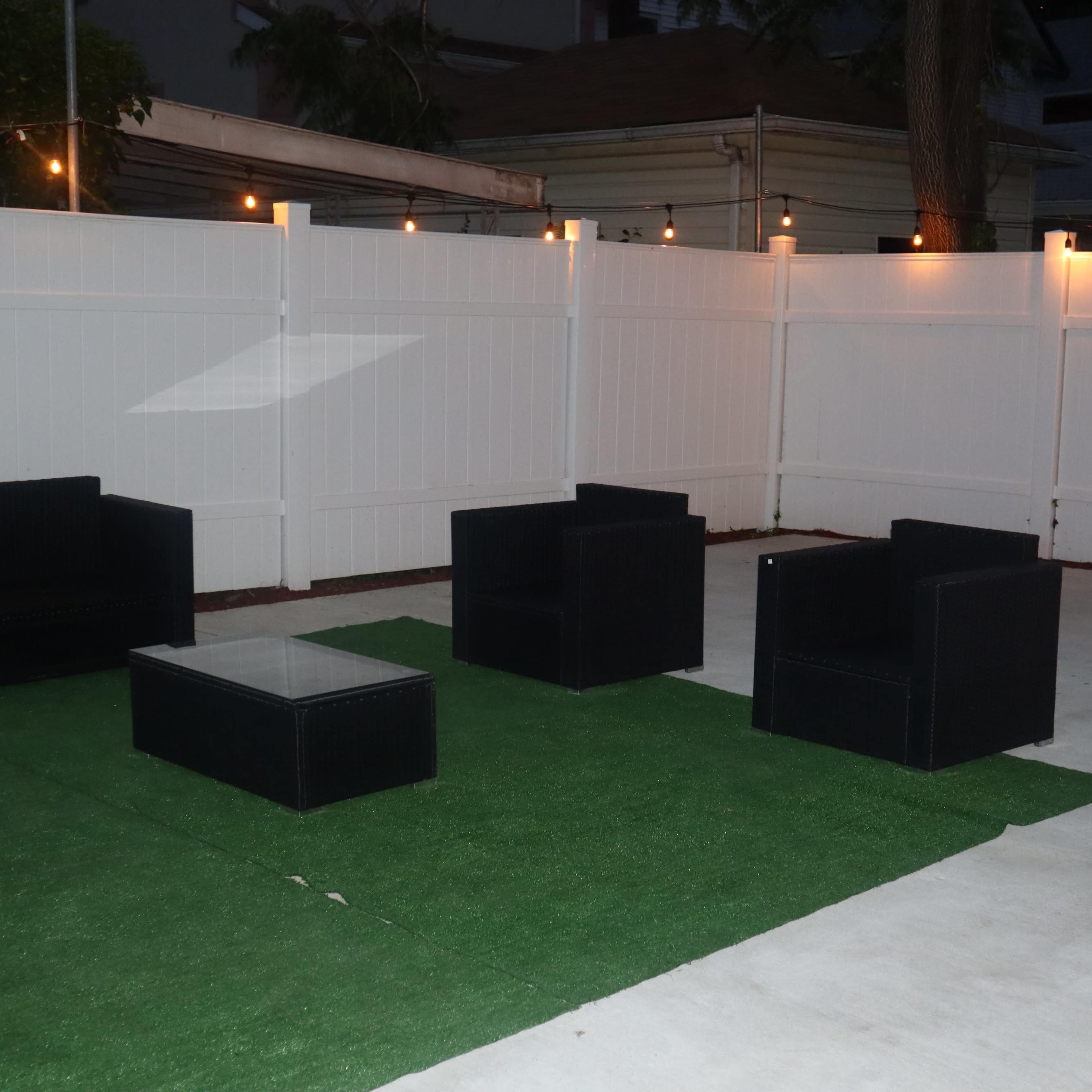 A patio area with black outdoor furniture arranged on synthetic grass, illuminated by string lights along a white fence.