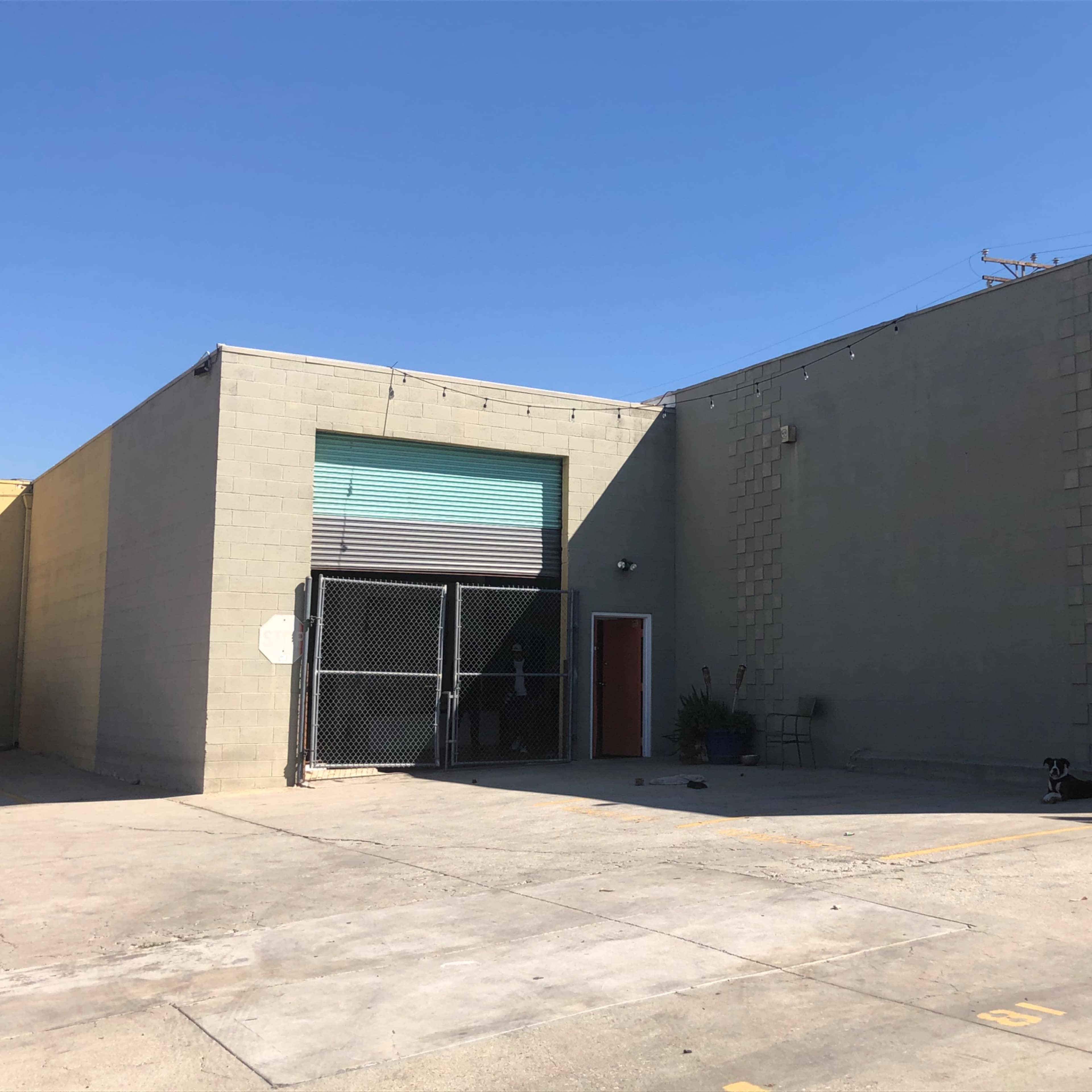 A gray industrial building with a fenced-off entrance and a dog lying in the parking lot.