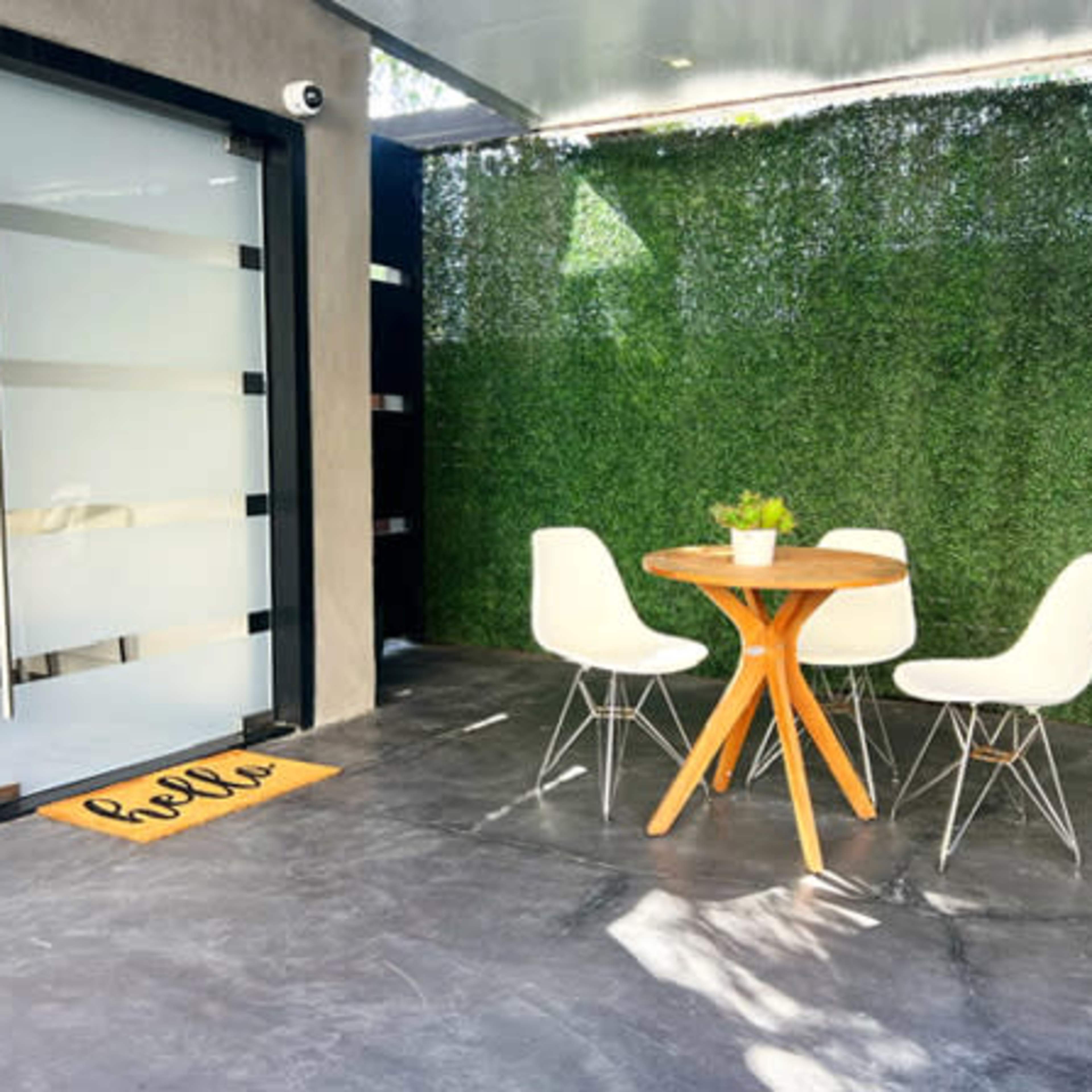 A round wooden table with three white chairs sits beside a green hedge wall, near a glass door with a welcome mat that says "hello."