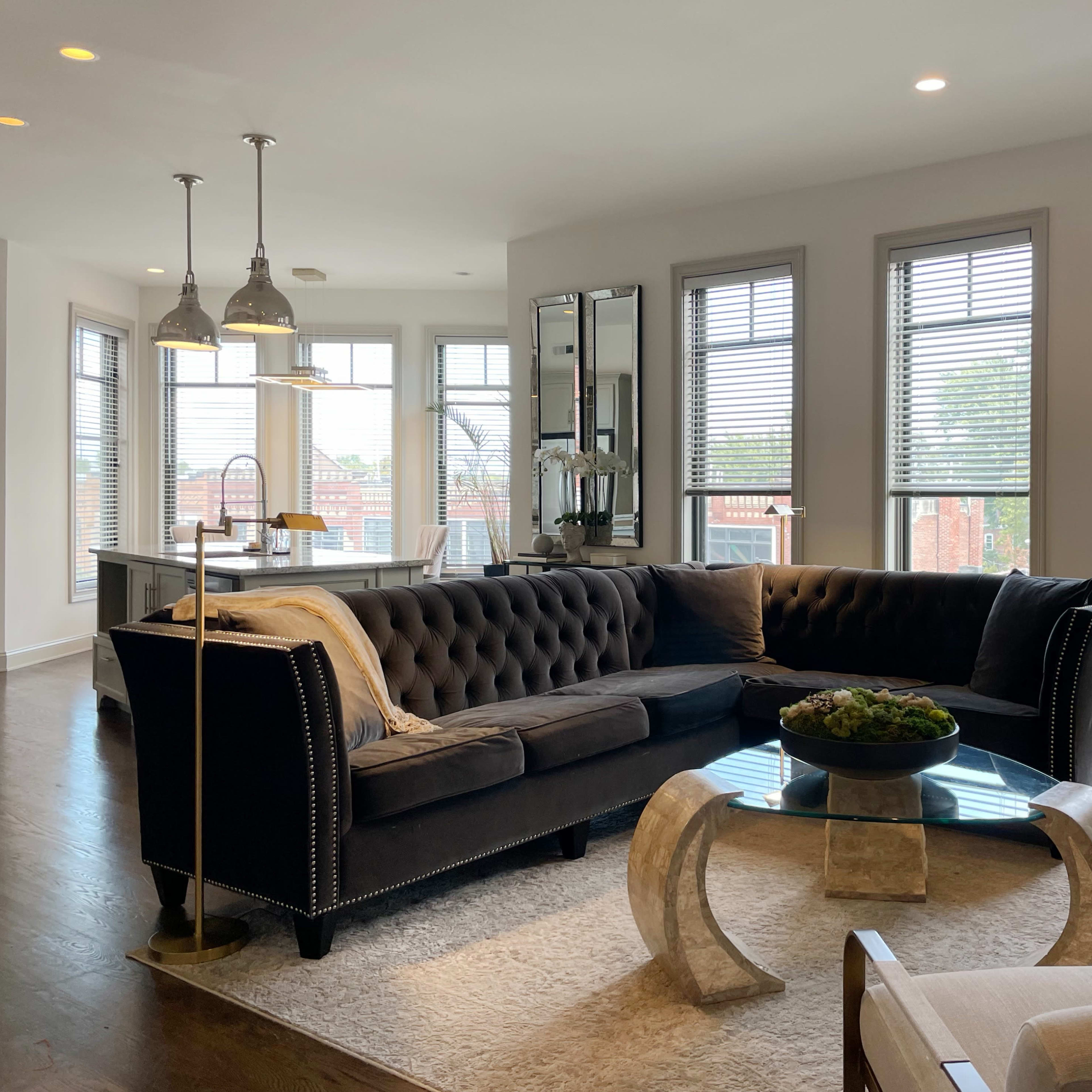 A modern living room with a large tufted sectional sofa, glass coffee table, and bright natural light streaming through multiple windows.