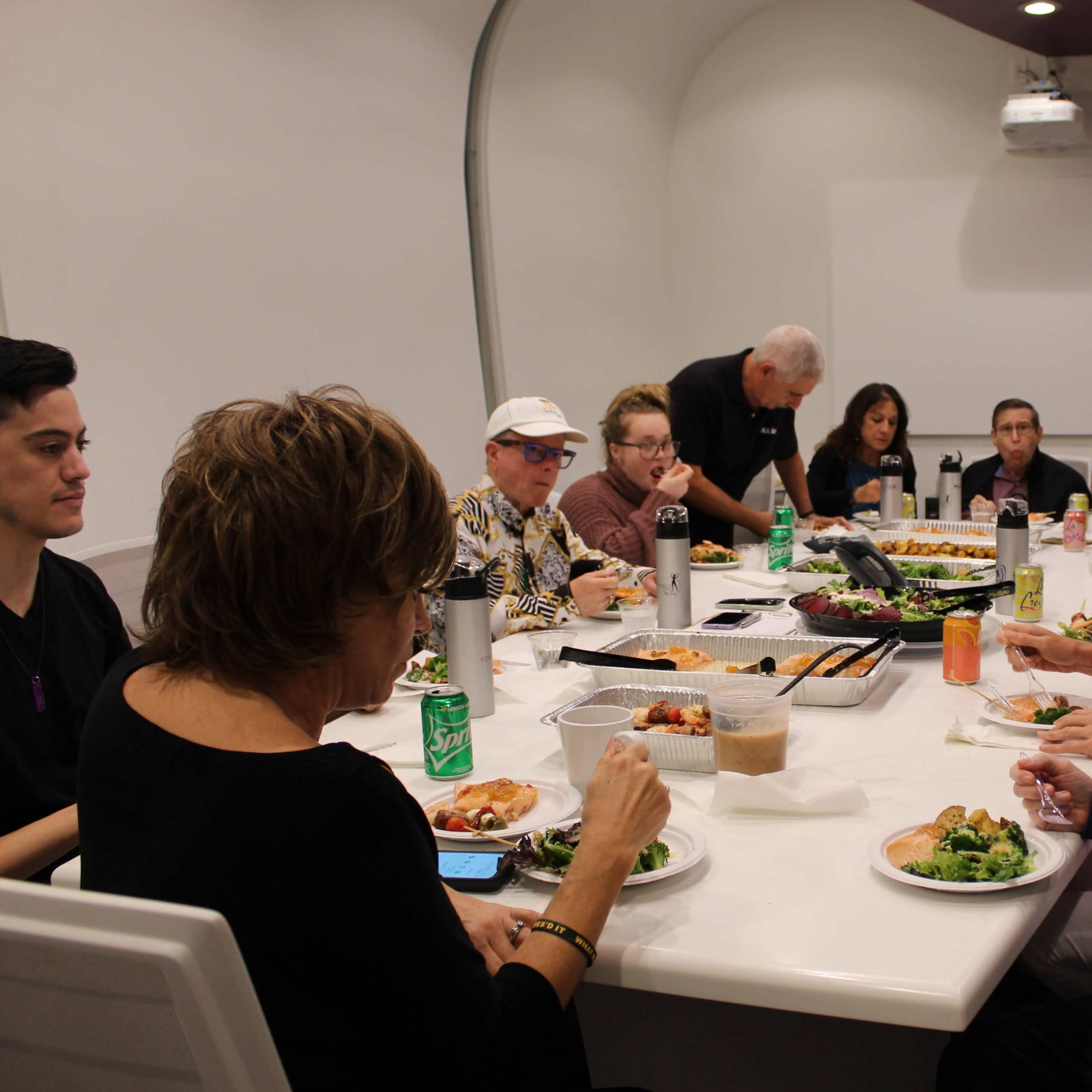 A group of people sits around a long table sharing a meal in a well-lit dining area.