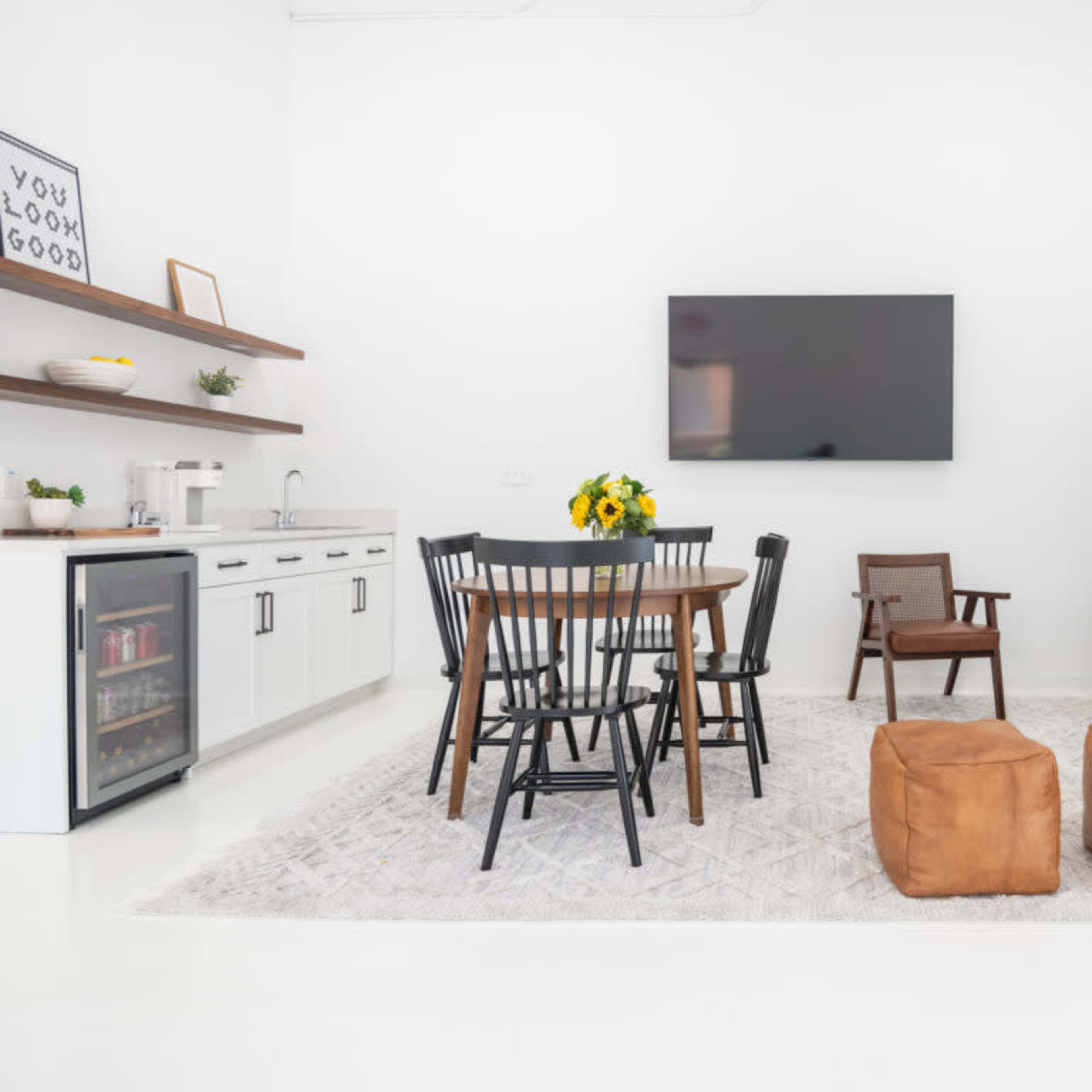 A modern, white-walled living space features a dining table with black chairs, a green sofa, a television on the wall, and a small kitchenette with a refrigerator.