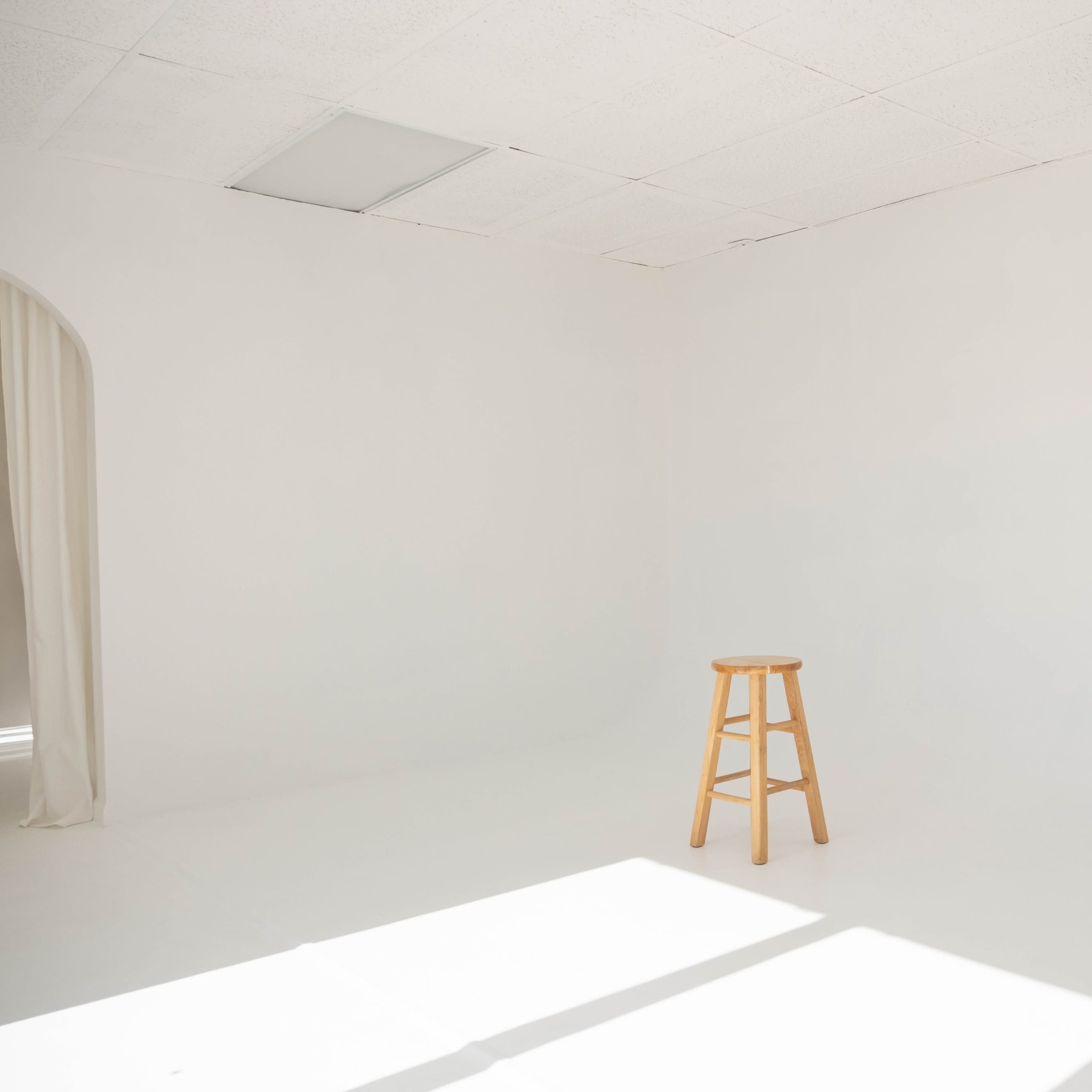 A wooden stool stands alone in the center of a bright, empty white room.