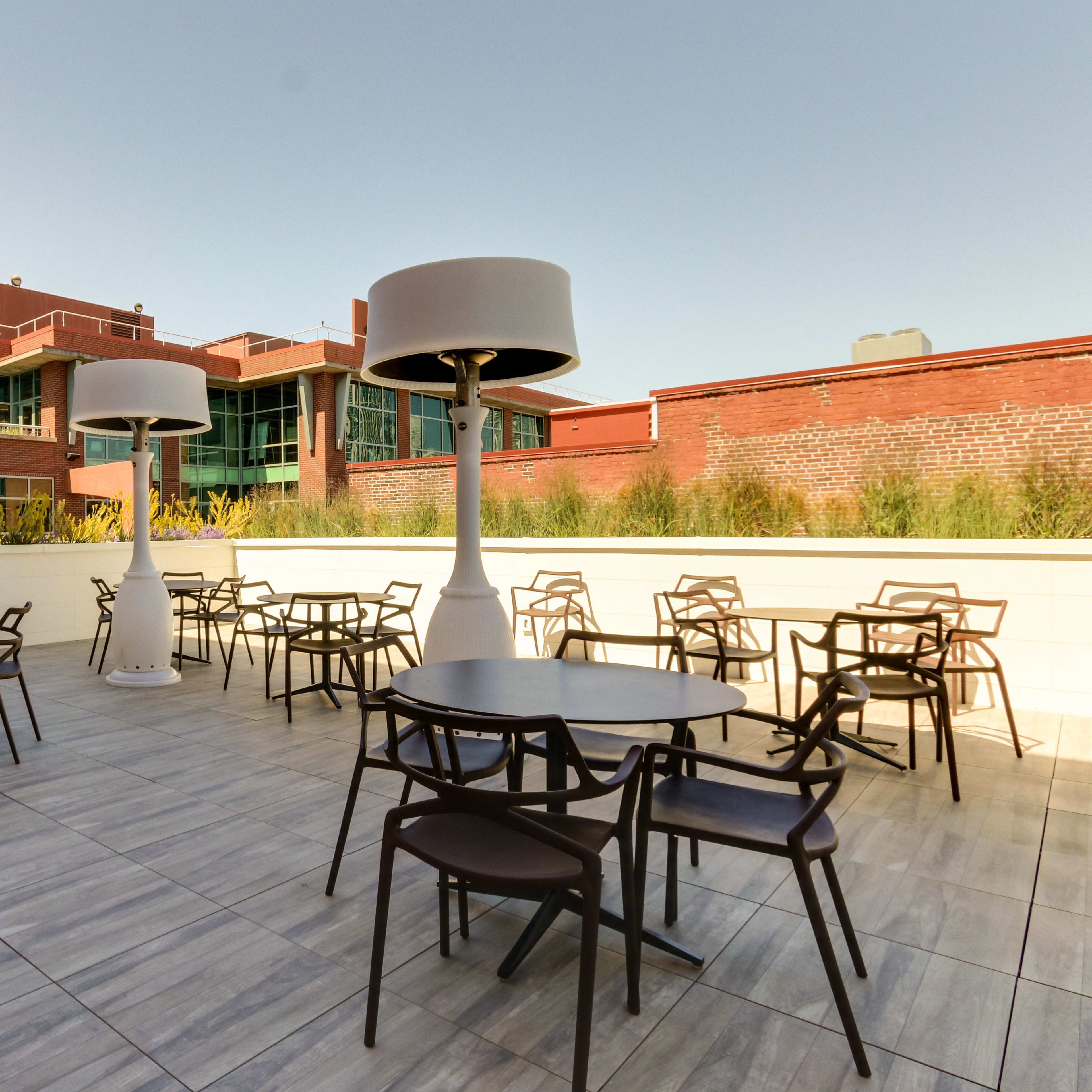 A spacious outdoor terrace featuring several round tables and oversized lamps, bordered by a building with large glass windows.