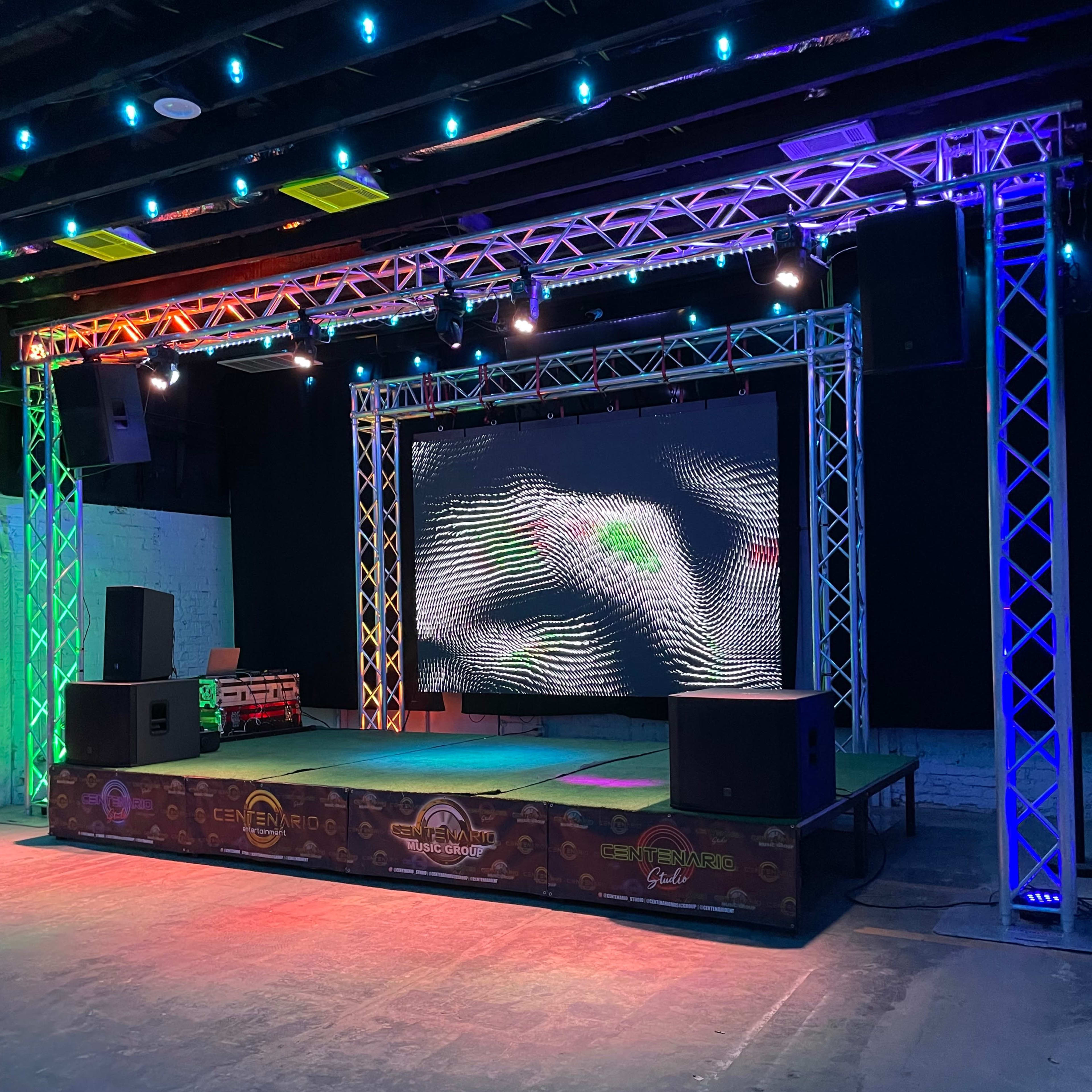A stage setup with colorful lighting, a large screen displaying wavy patterns, and speakers on either side, positioned in a spacious, dark venue.