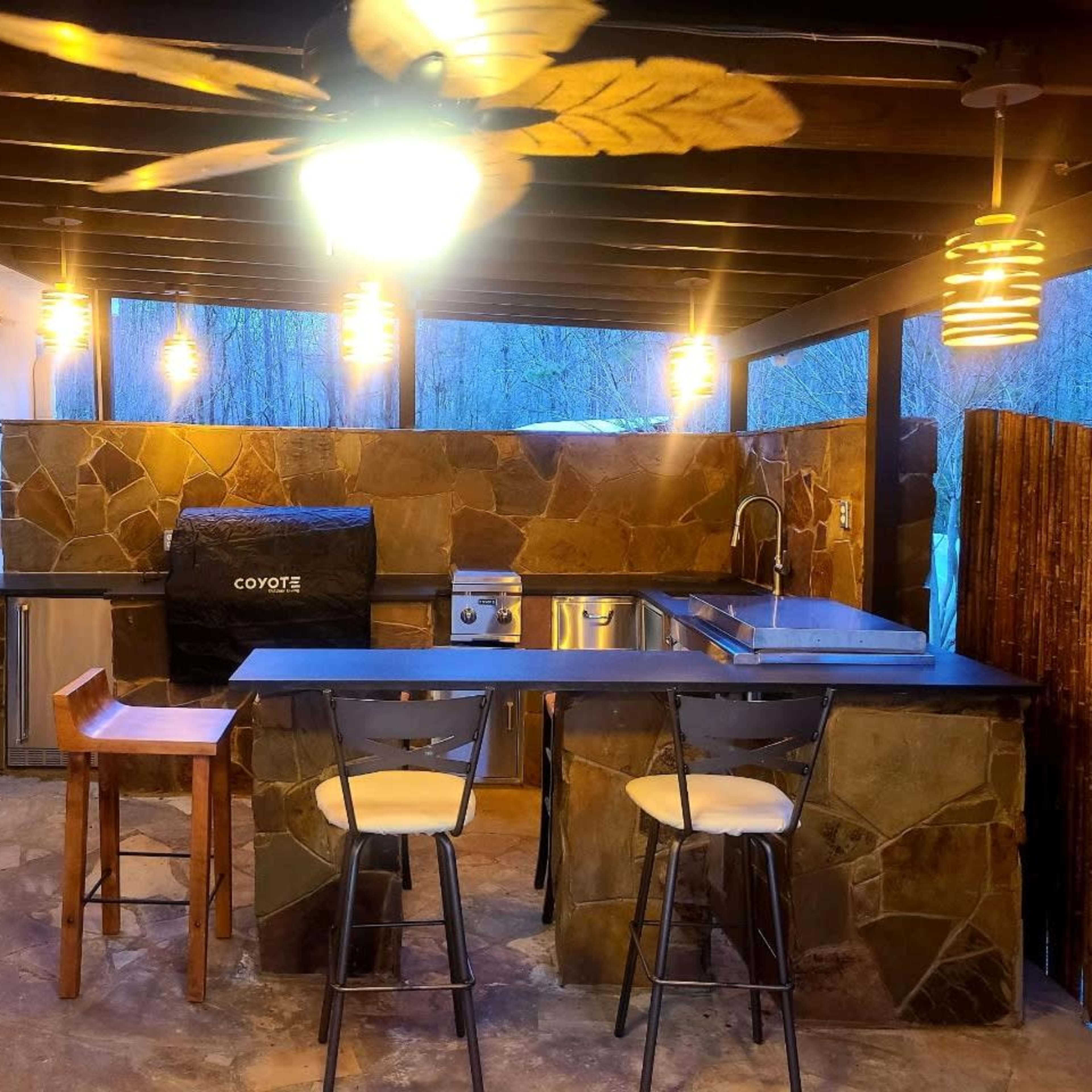 An outdoor kitchen with a stone countertop, a grill, and two high stools under a covered area illuminated by pendant lights.
