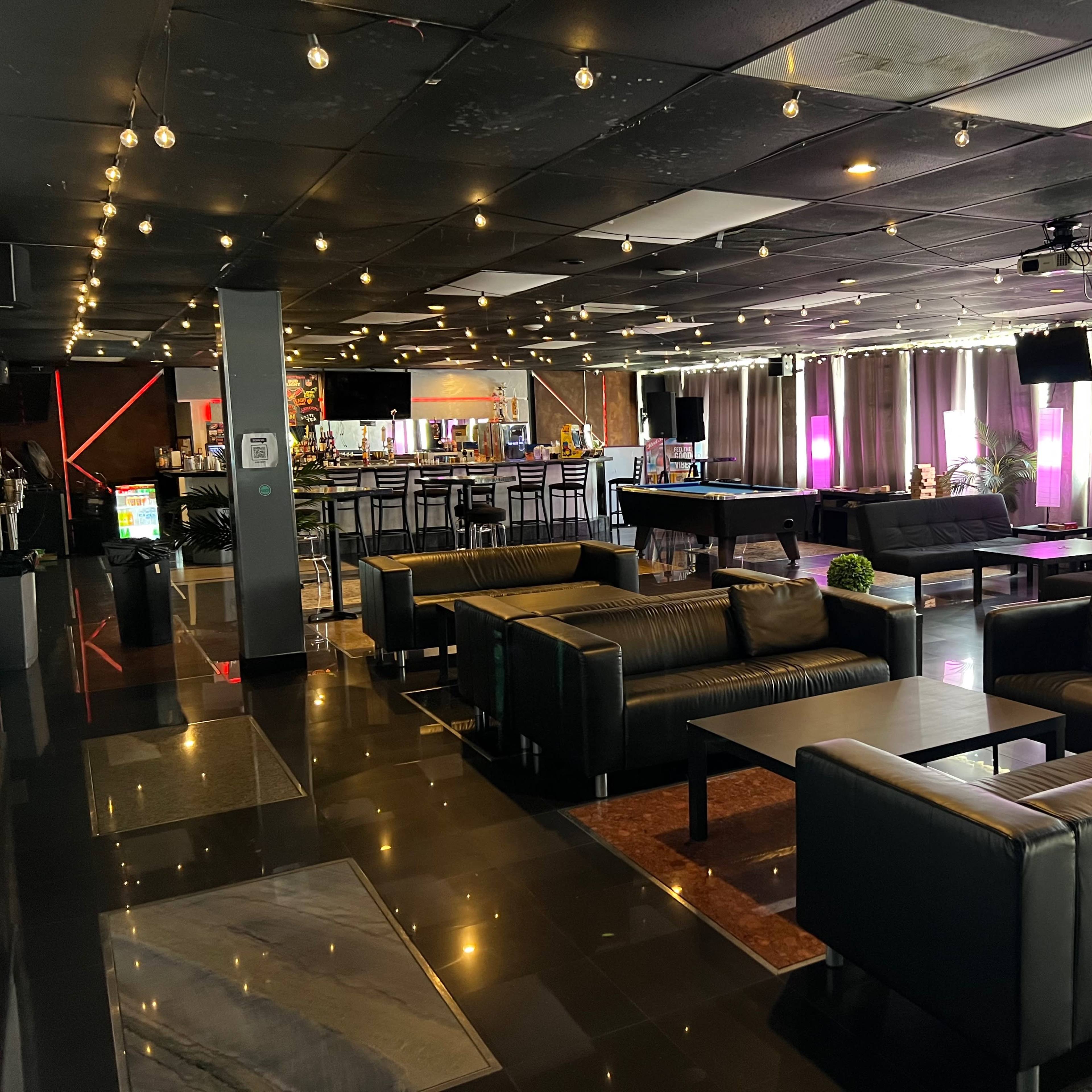 A modern lounge area with black leather seating, illuminated by soft lighting and decorated with plants in a spacious environment.