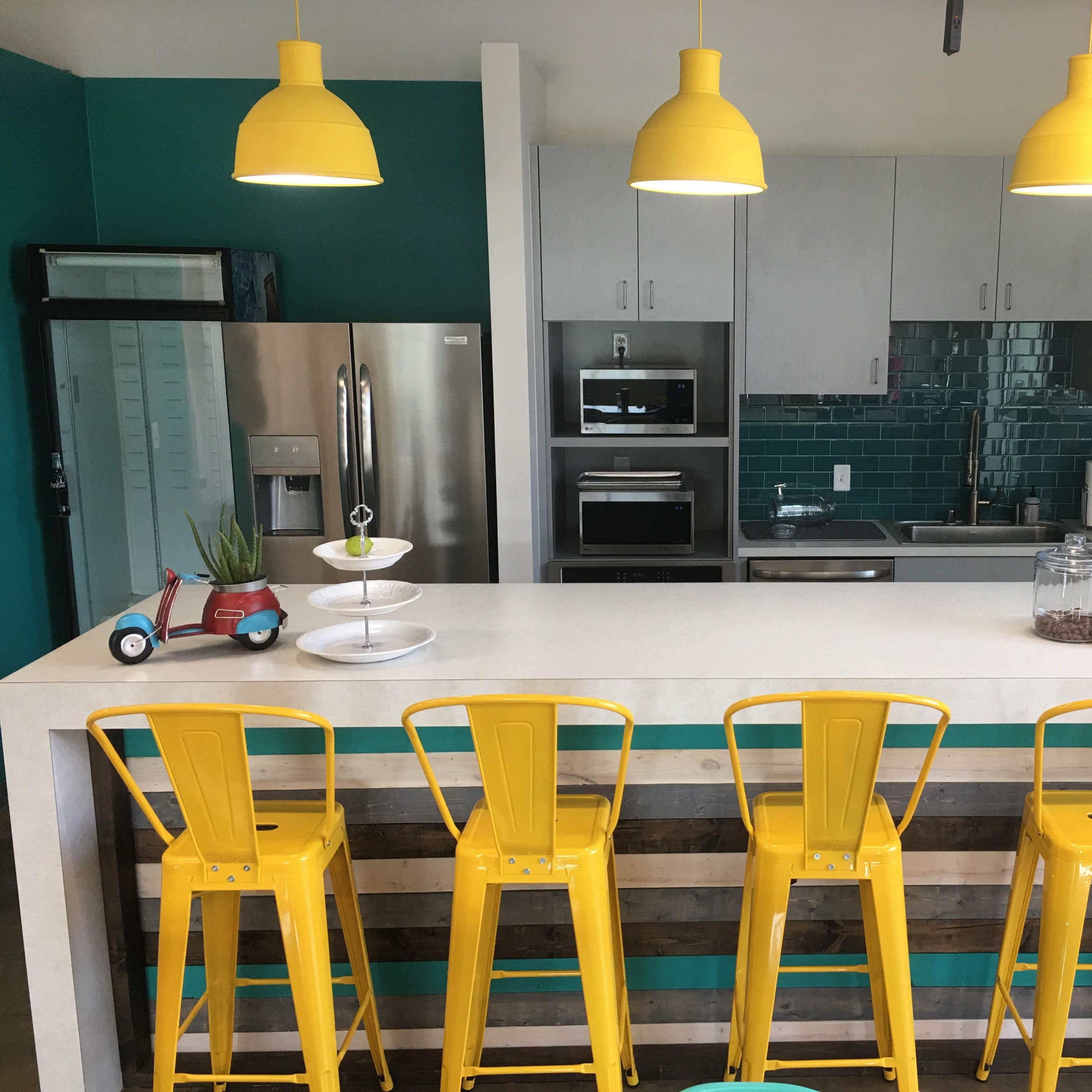 A modern kitchen features a large island with yellow bar stools, stainless steel appliances, and teal and gray cabinetry.