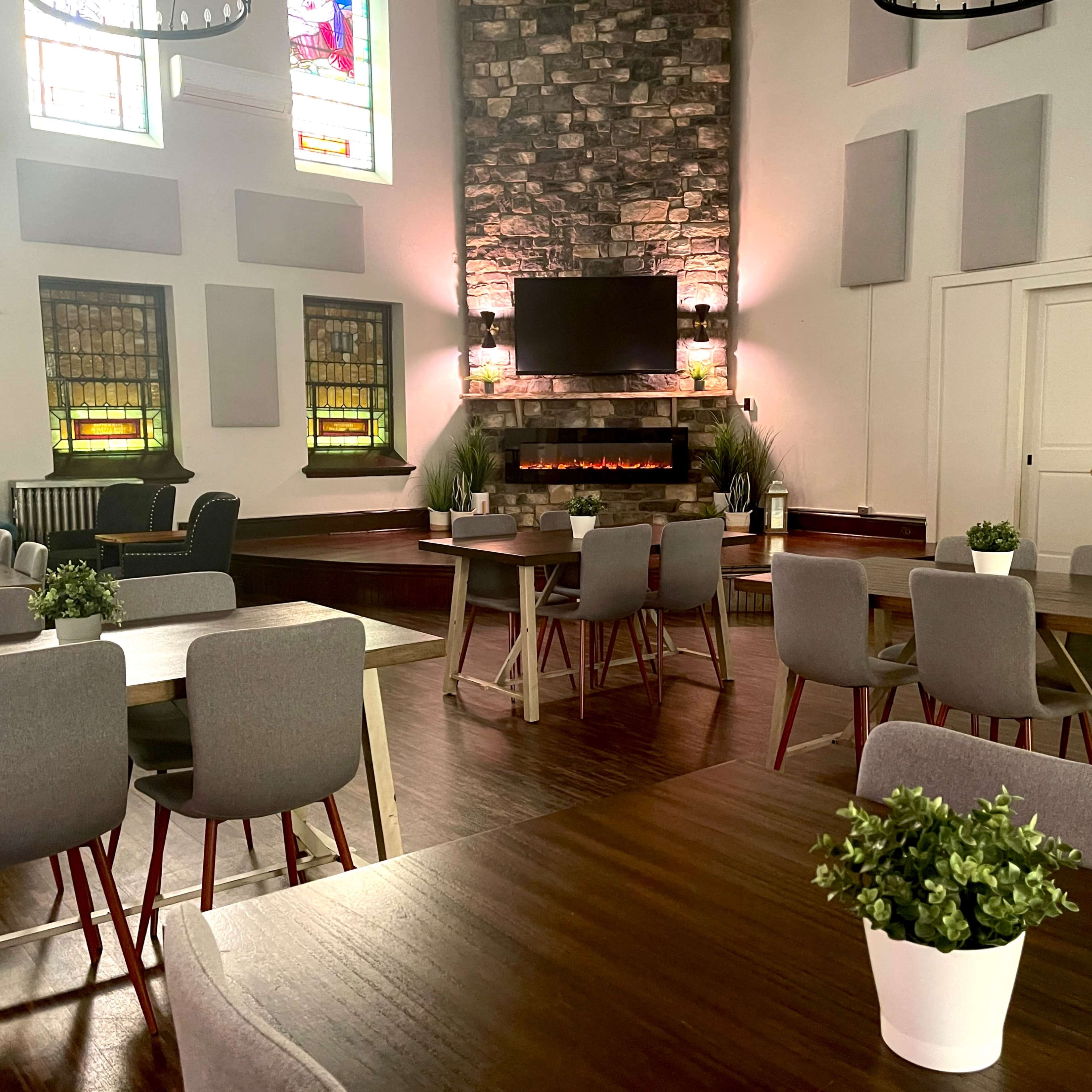 A spacious, well-lit room with multiple tables arranged around a modern fireplace and stained glass windows.
