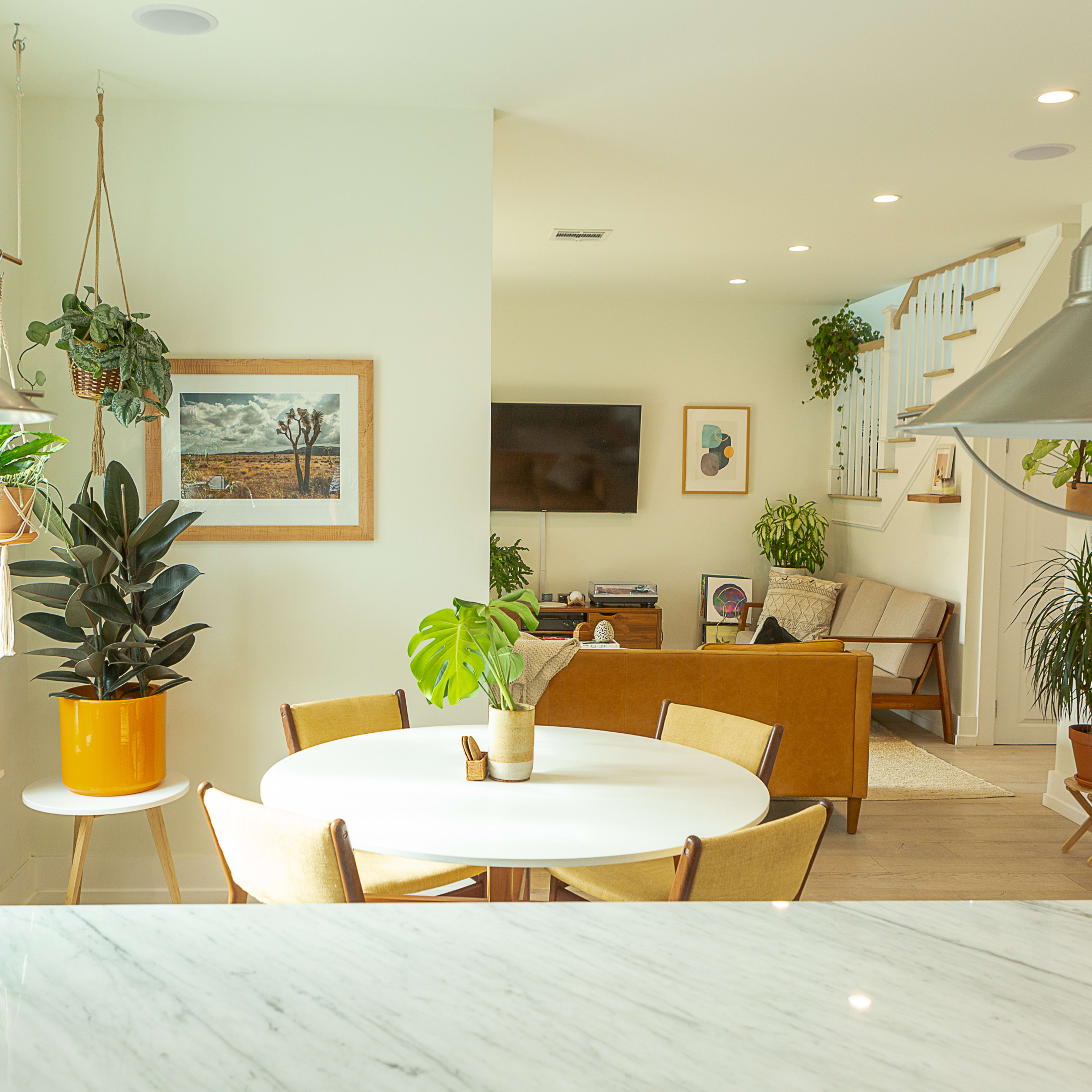 A bright, modern living space features a round dining table surrounded by chairs, with plants adorning the windows and a cozy seating area visible in the background.
