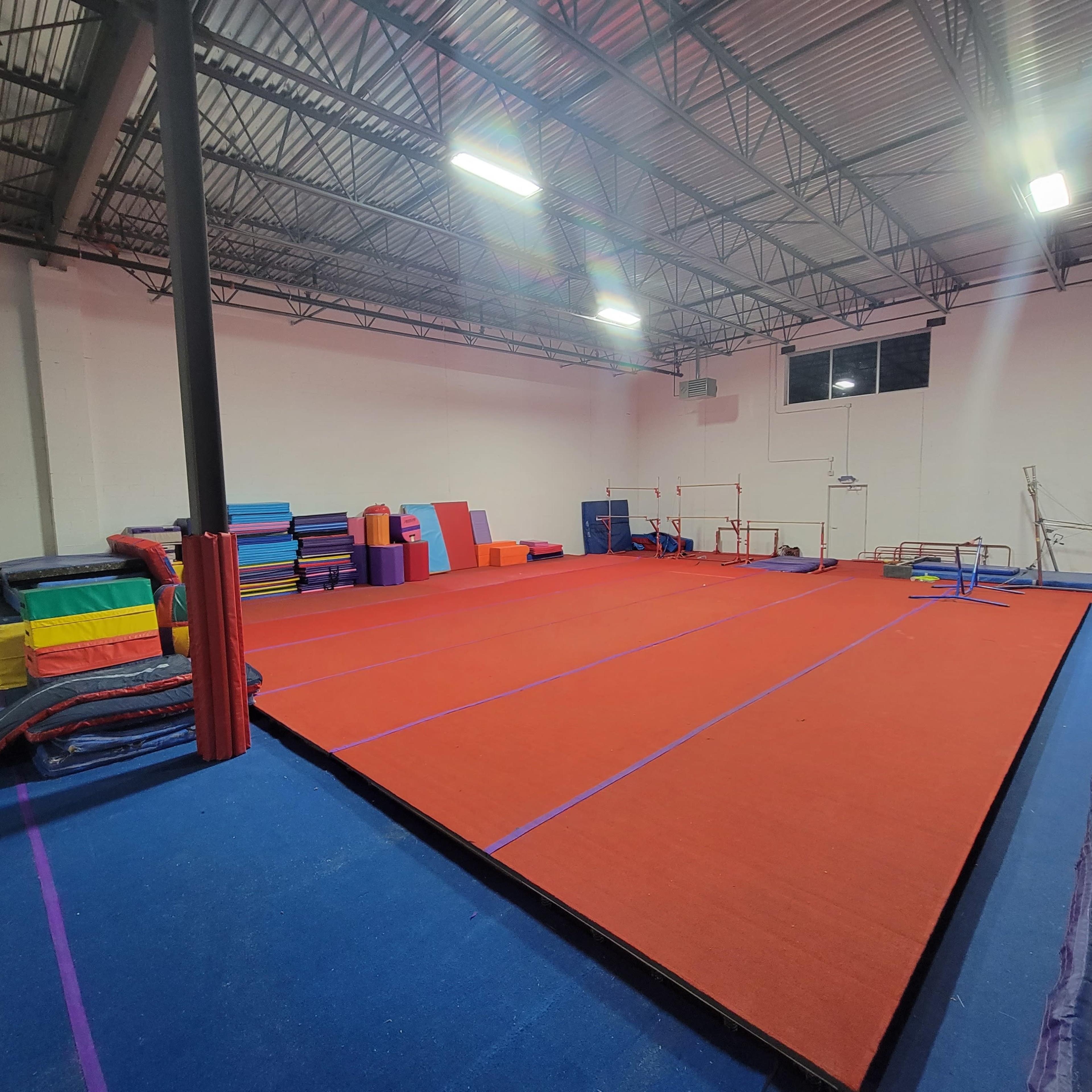 An empty gymnastics facility with red and blue mats arranged on the floor and various gymnastic equipment positioned along the walls.