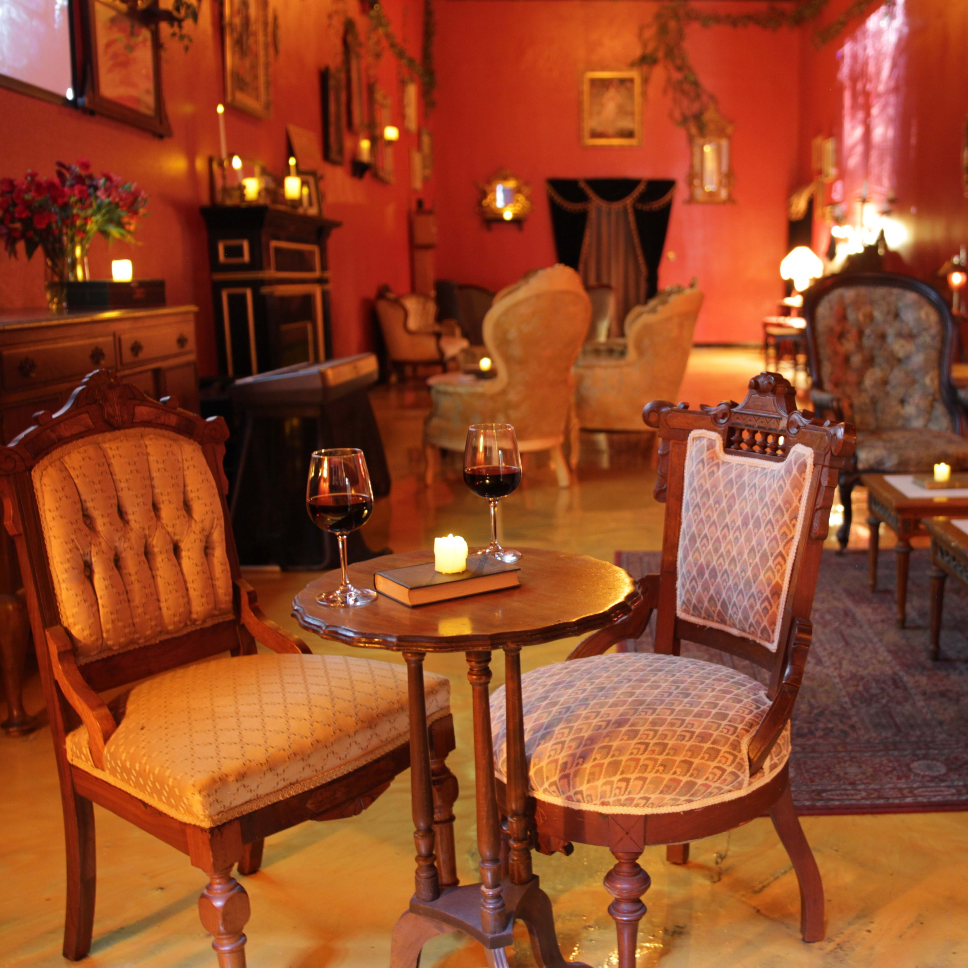 The scene features a cozy lounge area with vintage furniture, including two upholstered chairs, a small round table with glasses of red wine, and soft lighting from candles.