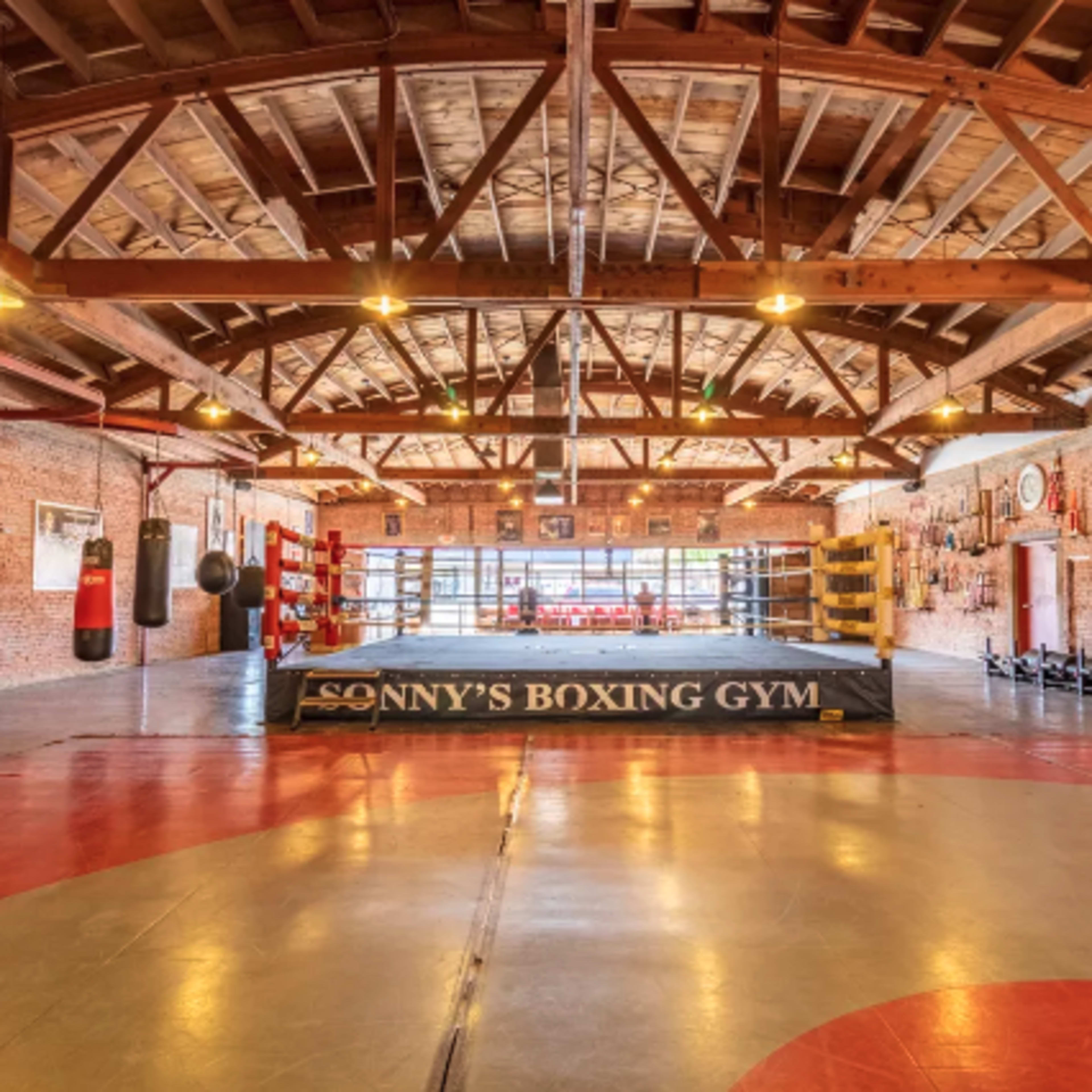 An empty boxing gym with a ring in the center, surrounded by various training equipment and a high wooden ceiling.