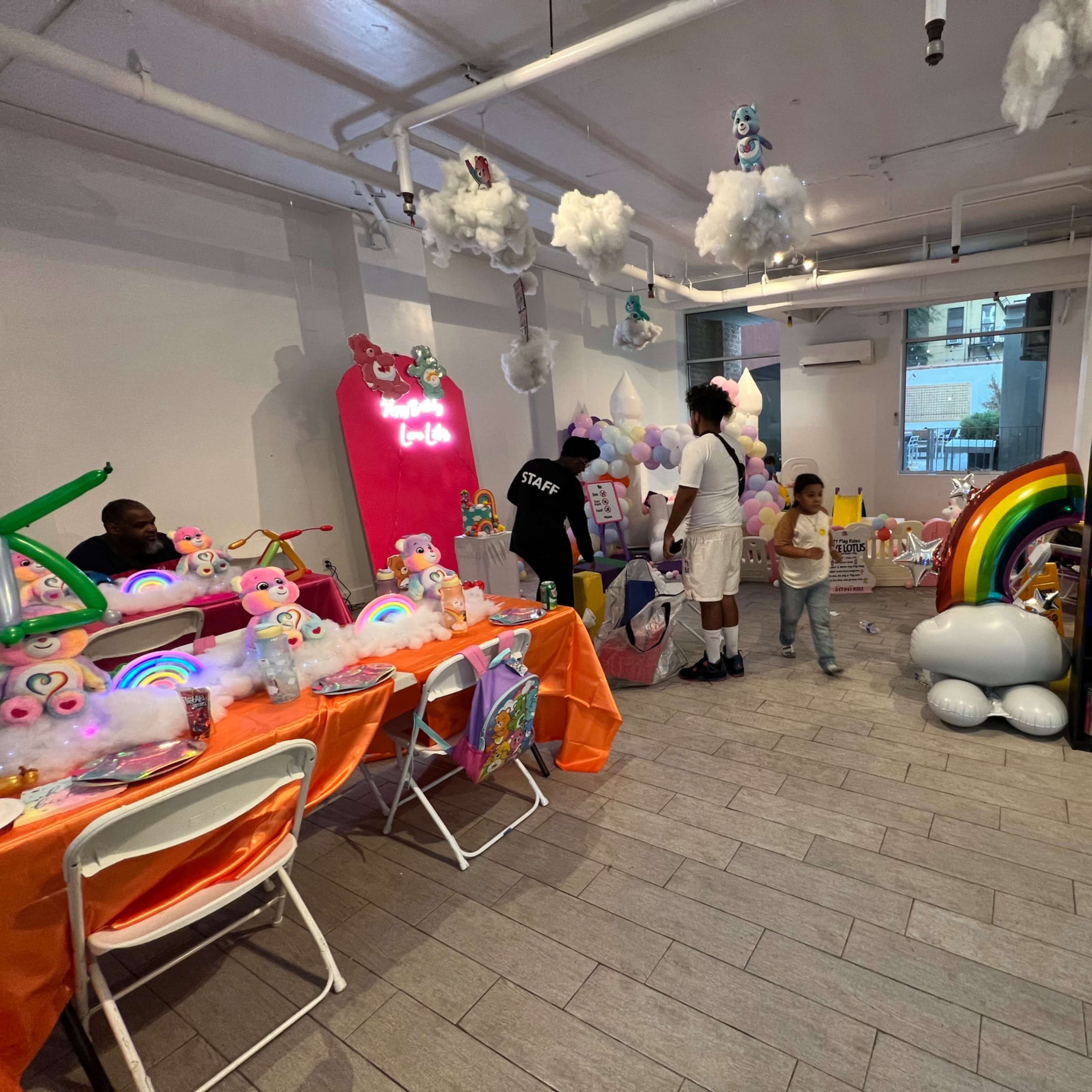 The scene shows a brightly decorated indoor space with colorful decorations, including staged toys, a rainbow display, and a mix of people engaging in activities.