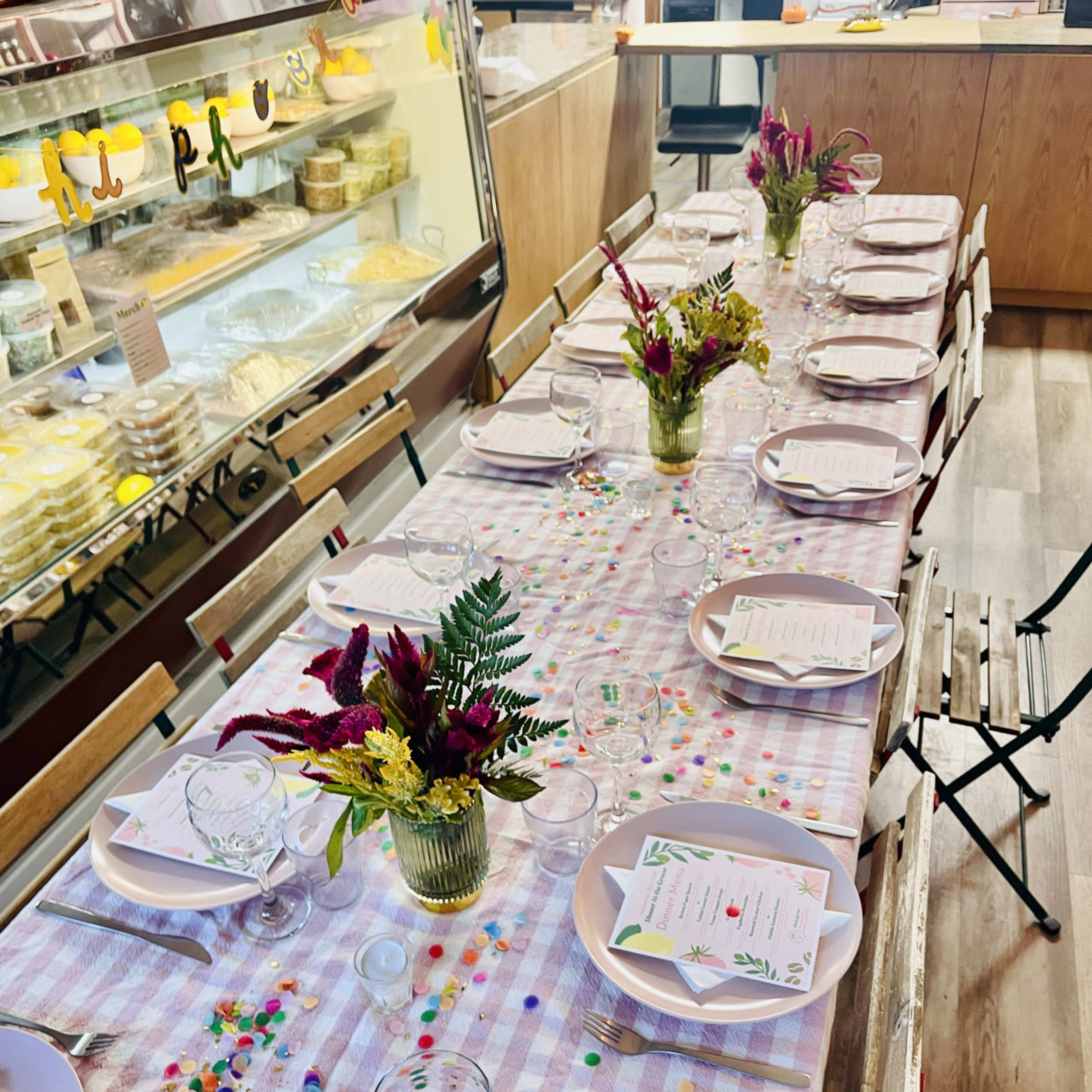 A long table is set with plates, glasses, and colorful flower arrangements, surrounded by chairs in a brightly lit space with a display of food in the background.
