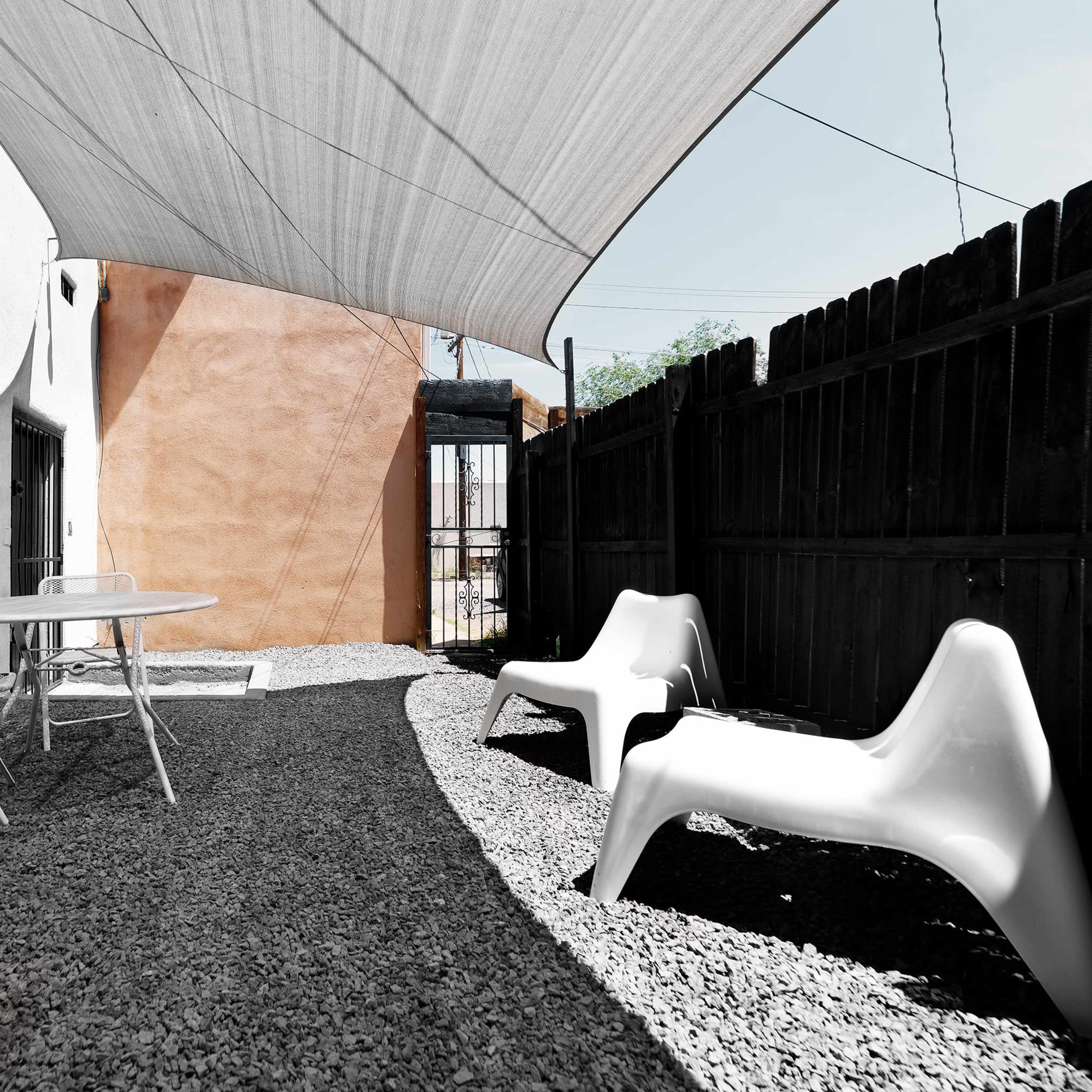 A shaded outdoor area with a gravel floor, featuring a white table and two modern chairs against a backdrop of a wooden fence and a stucco wall.