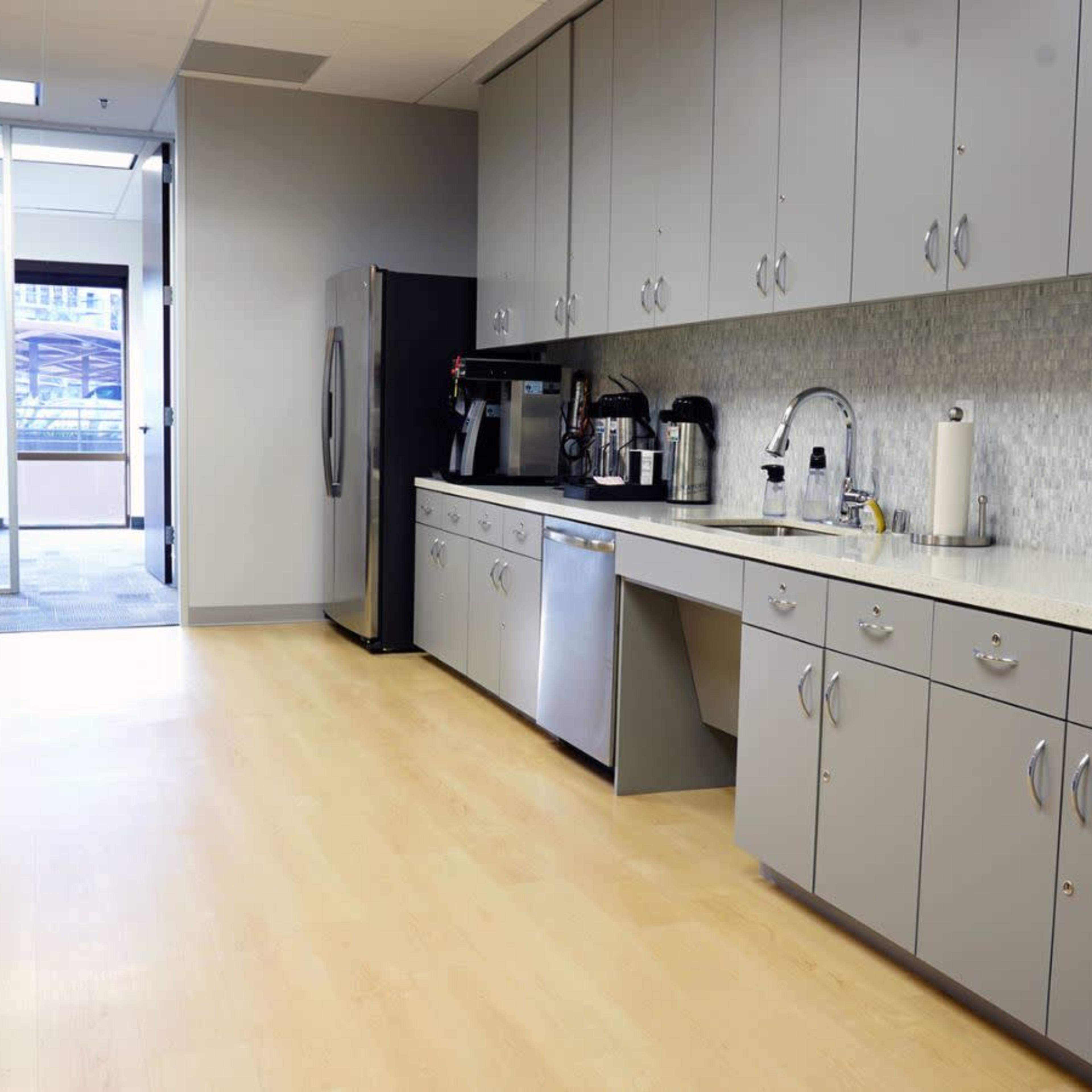 A modern kitchen area with gray cabinetry, a coffee machine, and appliances, featuring a large counter and an open door leading to an outdoor space.