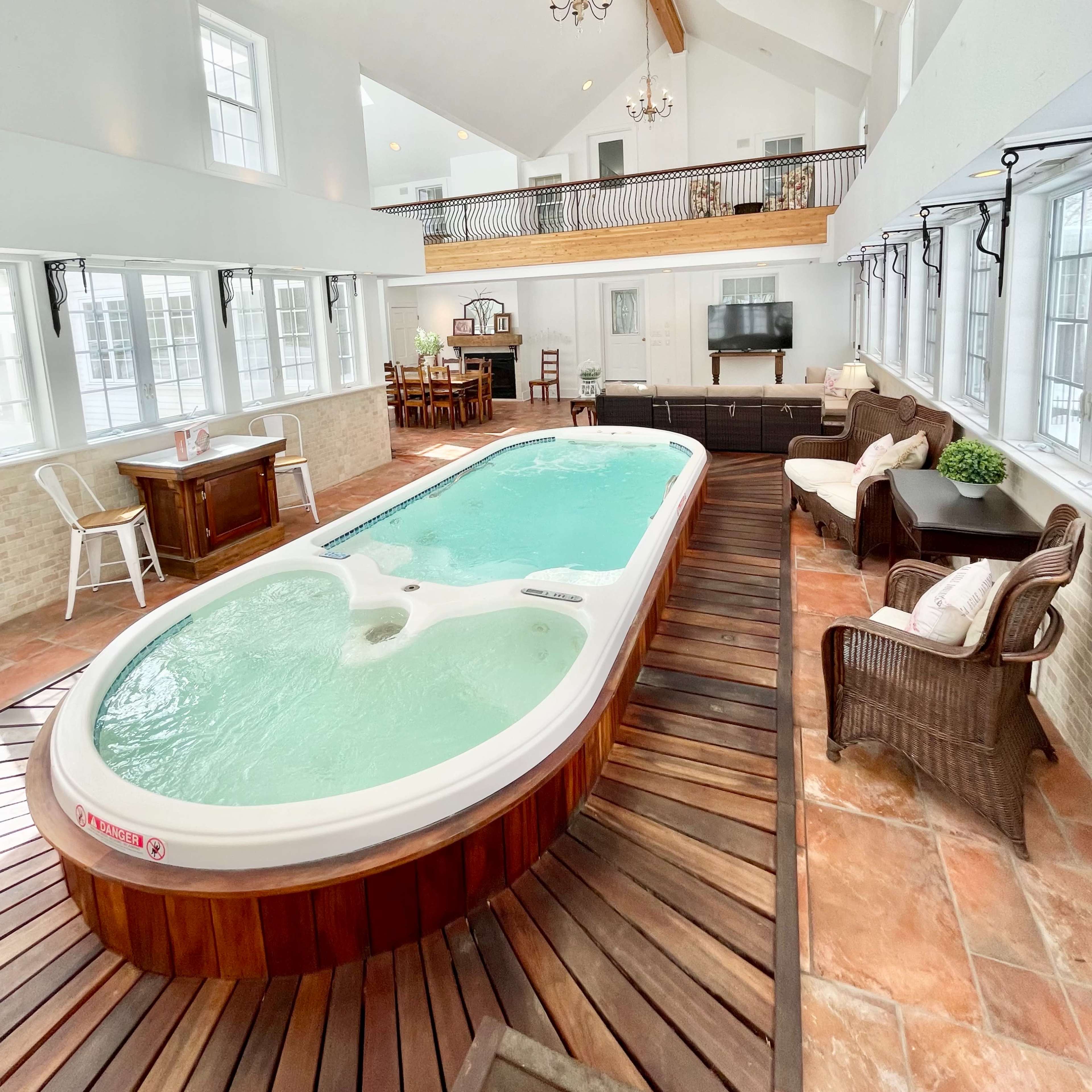 A spacious indoor area featuring a rectangular swimming pool surrounded by wooden decking, with a sitting area and dining space in the background.