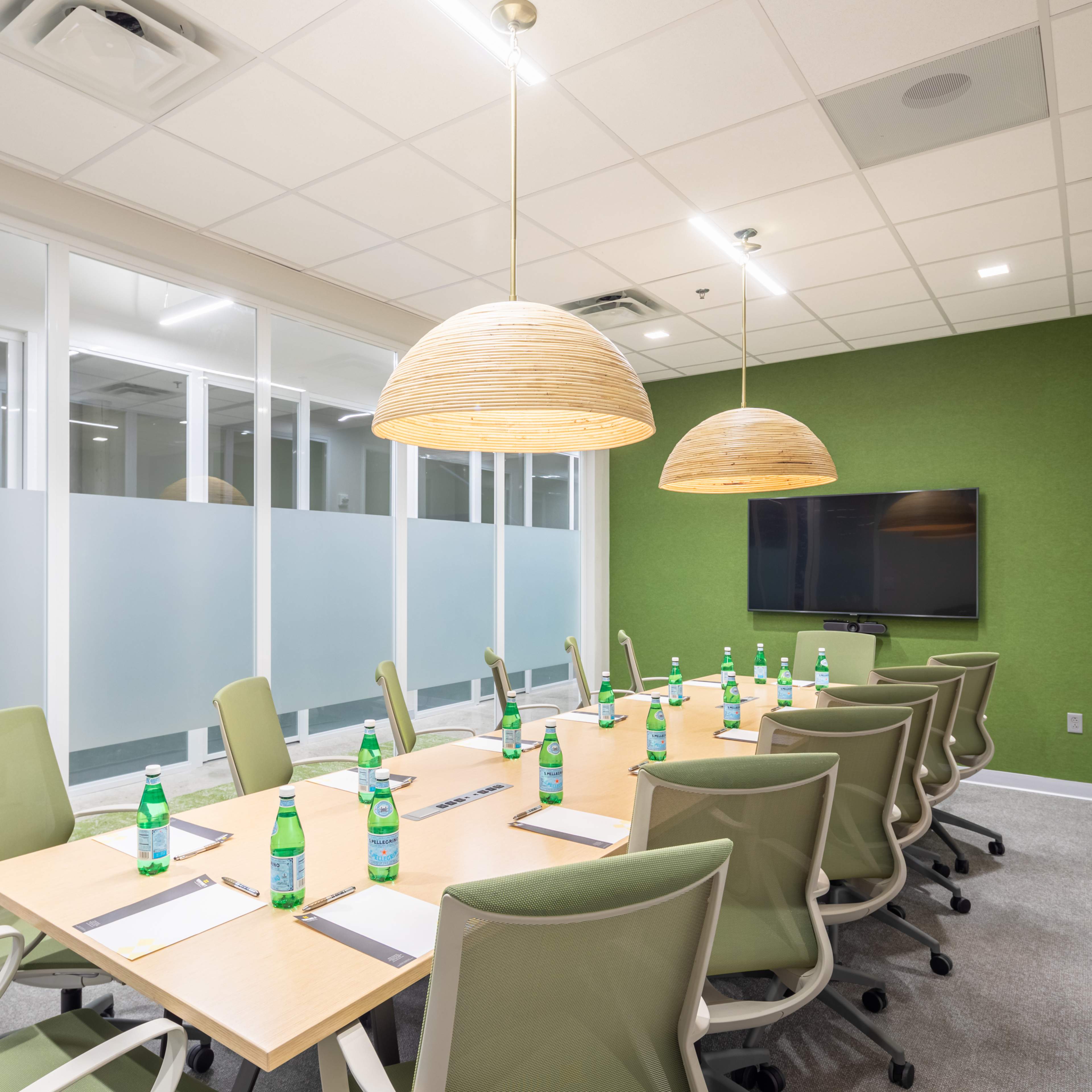 A modern conference room features a long table set with notepads and water bottles, surrounded by green chairs and illuminated by pendant lights.