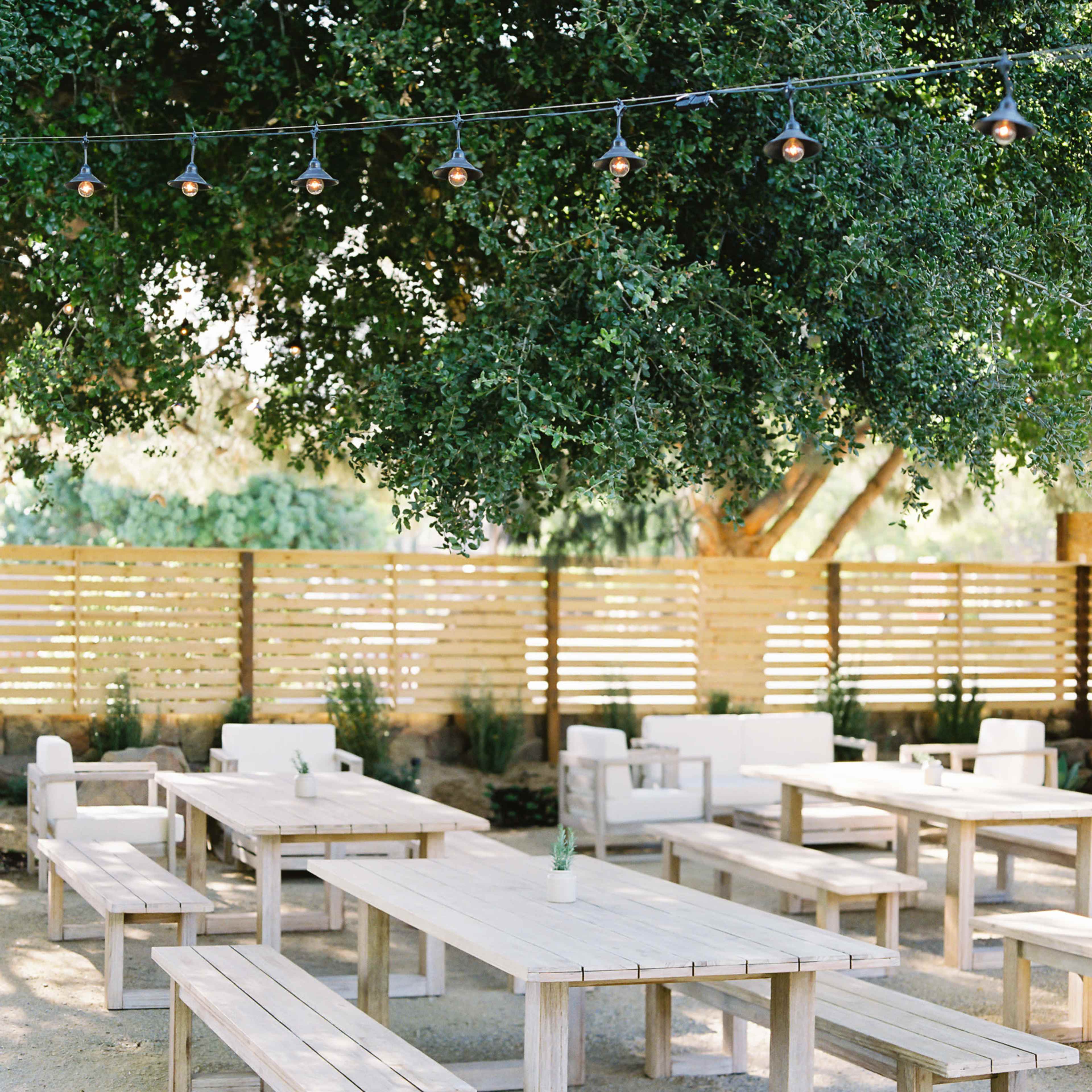 A selection of wooden tables and benches is arranged under a large tree, with string lights overhead and a wooden fence in the background.