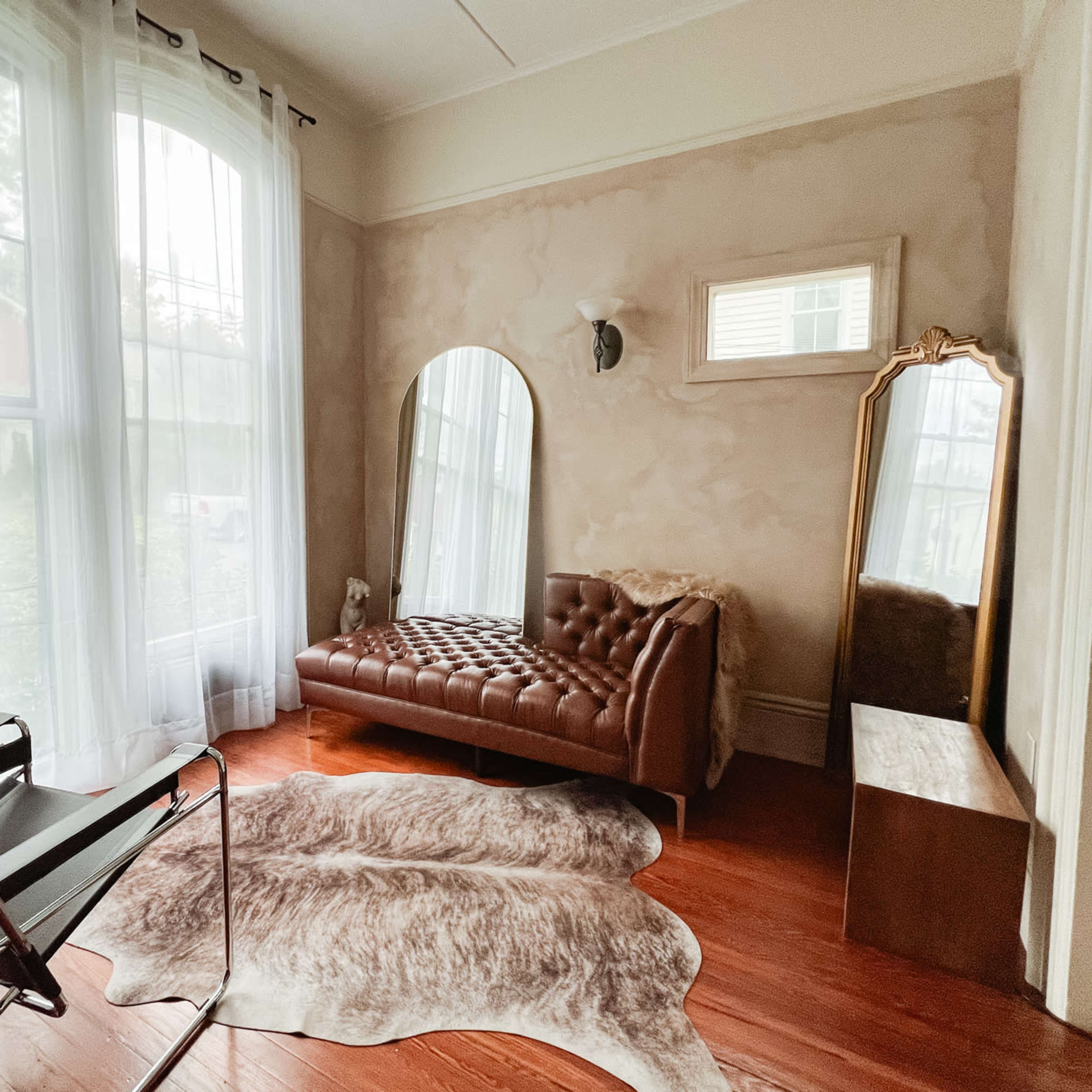 A corner of a room featuring a brown leather tufted sofa, a large mirror, a chair, and a cowhide rug on wooden flooring beside tall windows with sheer curtains.