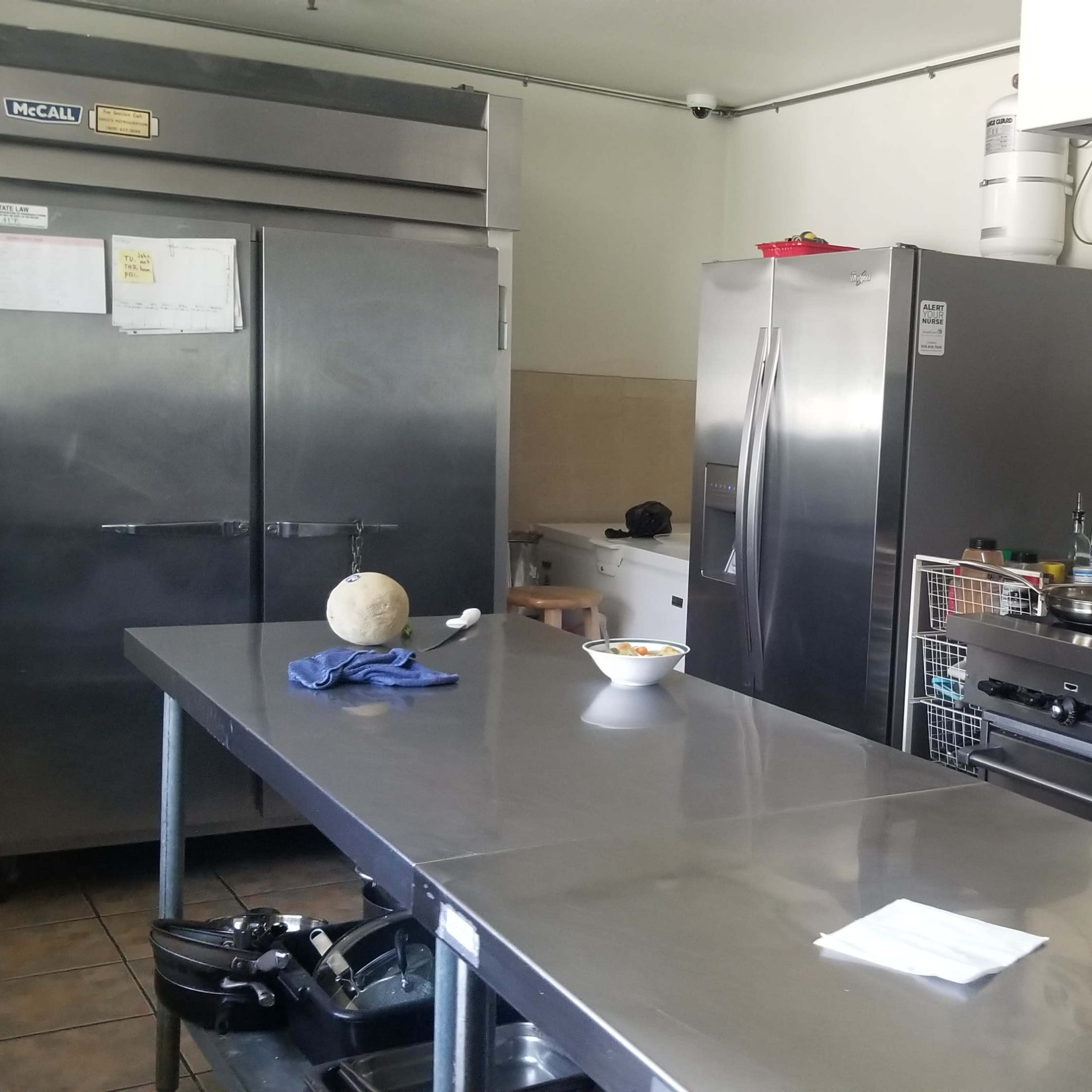 A commercial kitchen with stainless steel appliances, including a refrigerator and a range, featuring a countertop with a bowl of food and a kitchen towel.