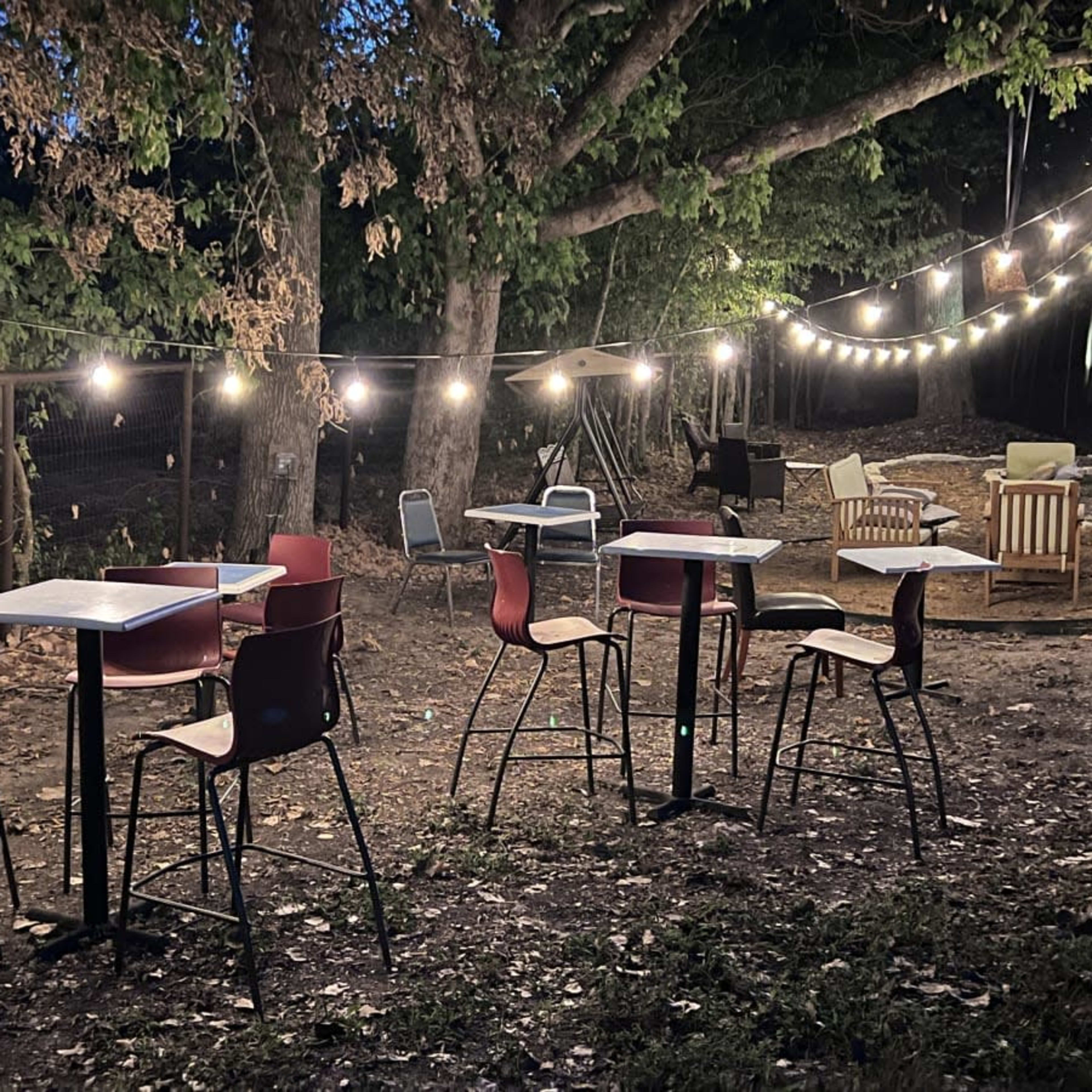 A small outdoor seating area with several tables and chairs is illuminated by string lights in a wooded setting.