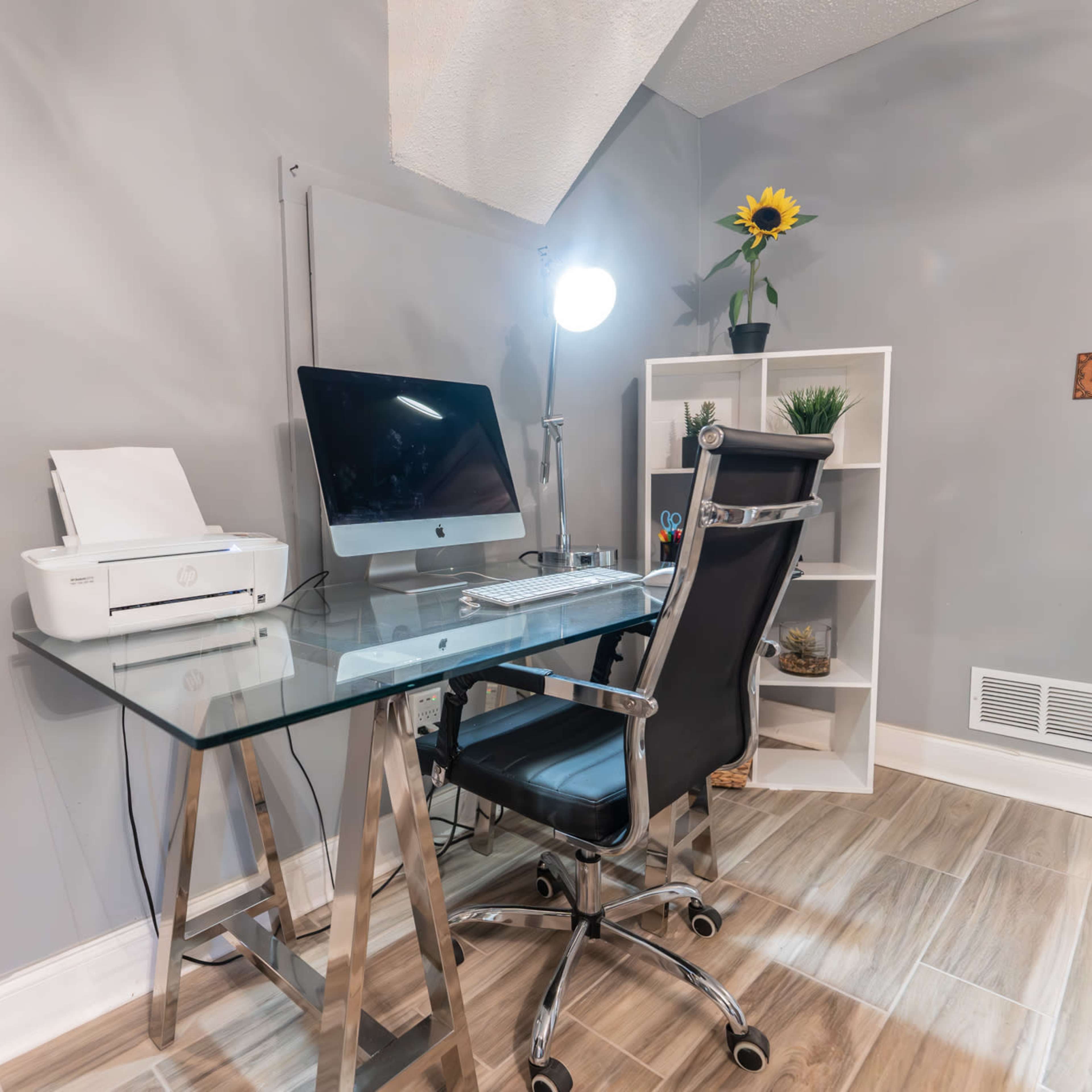 A modern home office setup featuring a glass desk with an iMac, a printer, a black ergonomic chair, a desk lamp, and a small white shelf with plants and a sunflower.