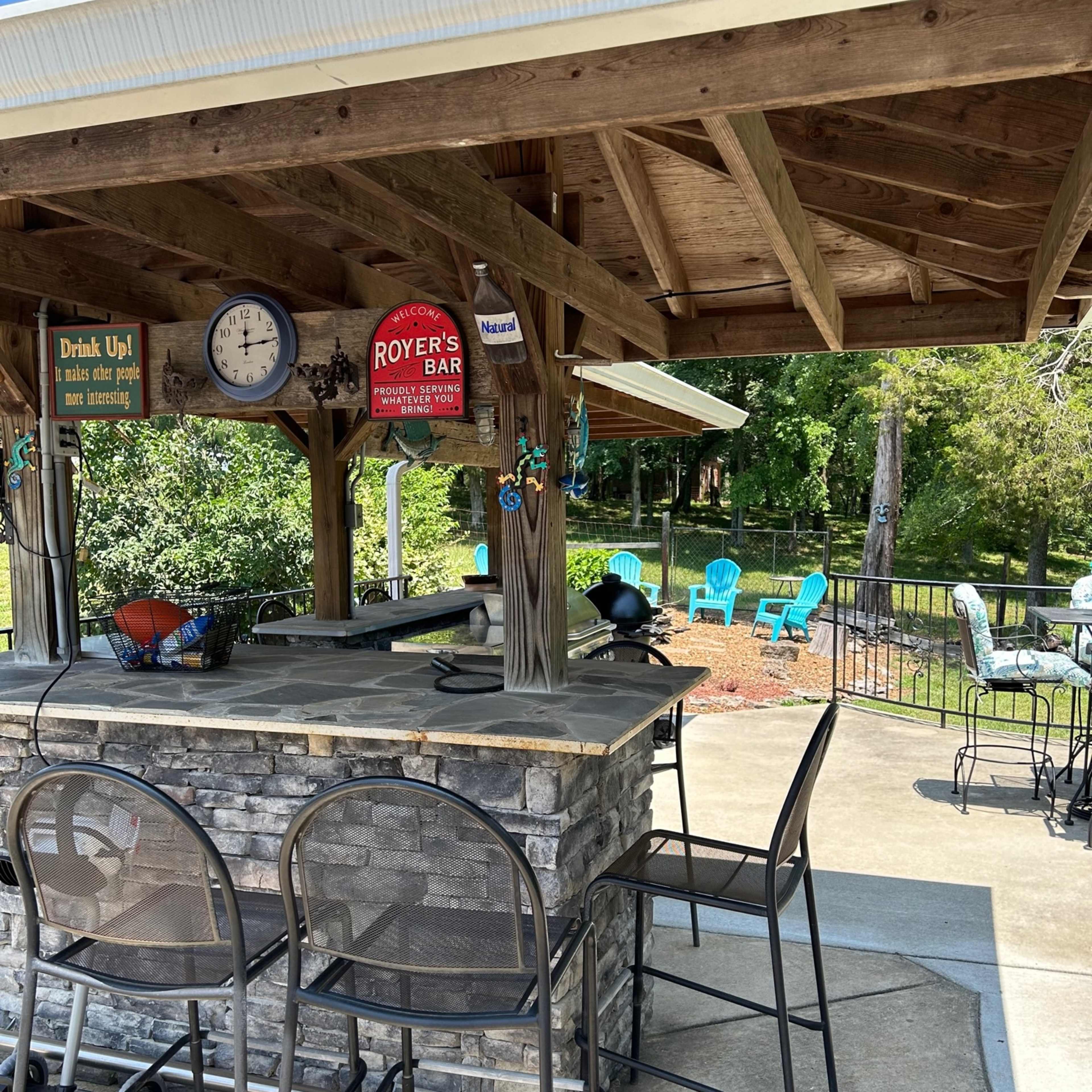 A stone bar with high chairs is situated under a wooden pavilion, surrounded by outdoor seating and greenery.