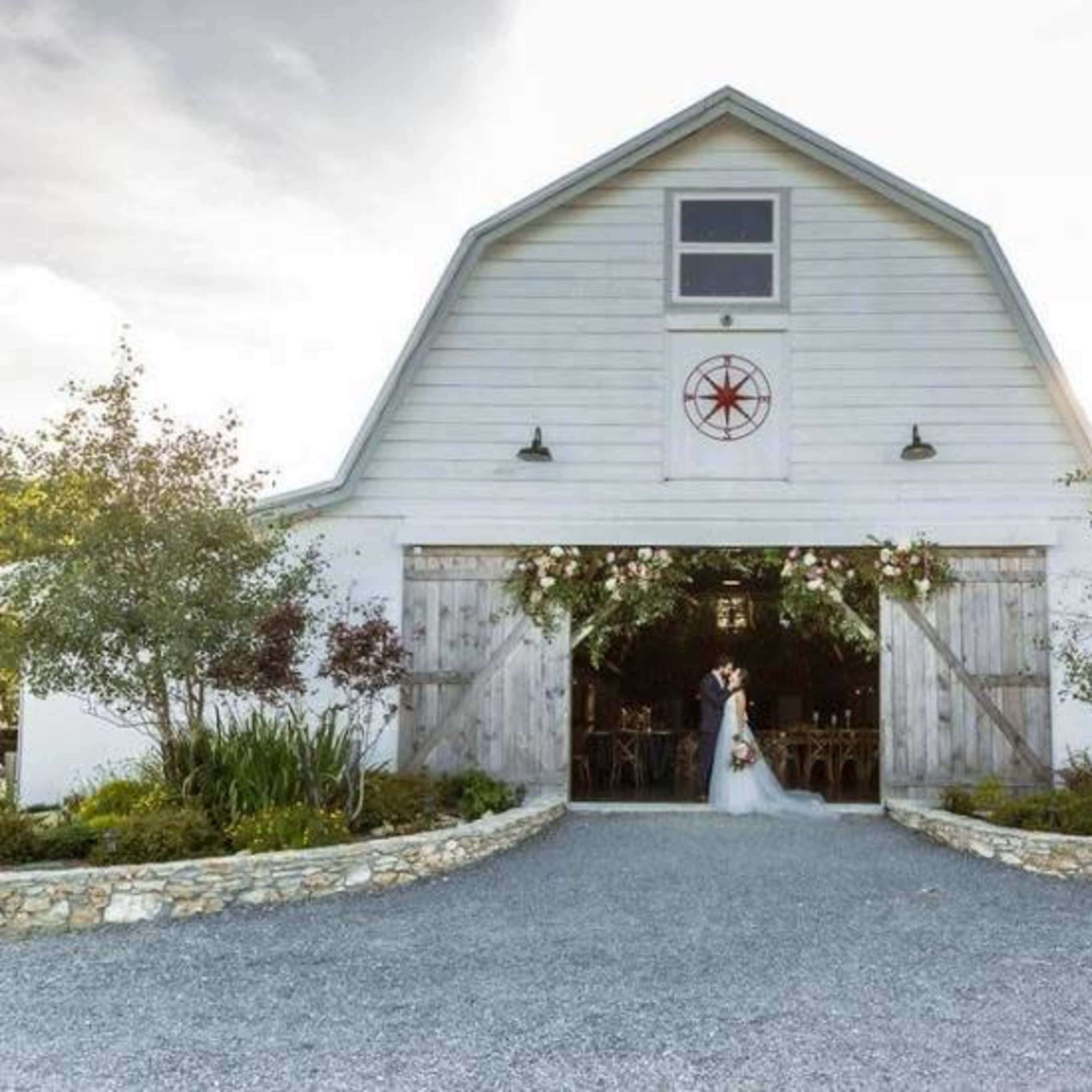 A bride and groom stand in front of a white barn with wooden doors adorned with flowers and greenery.