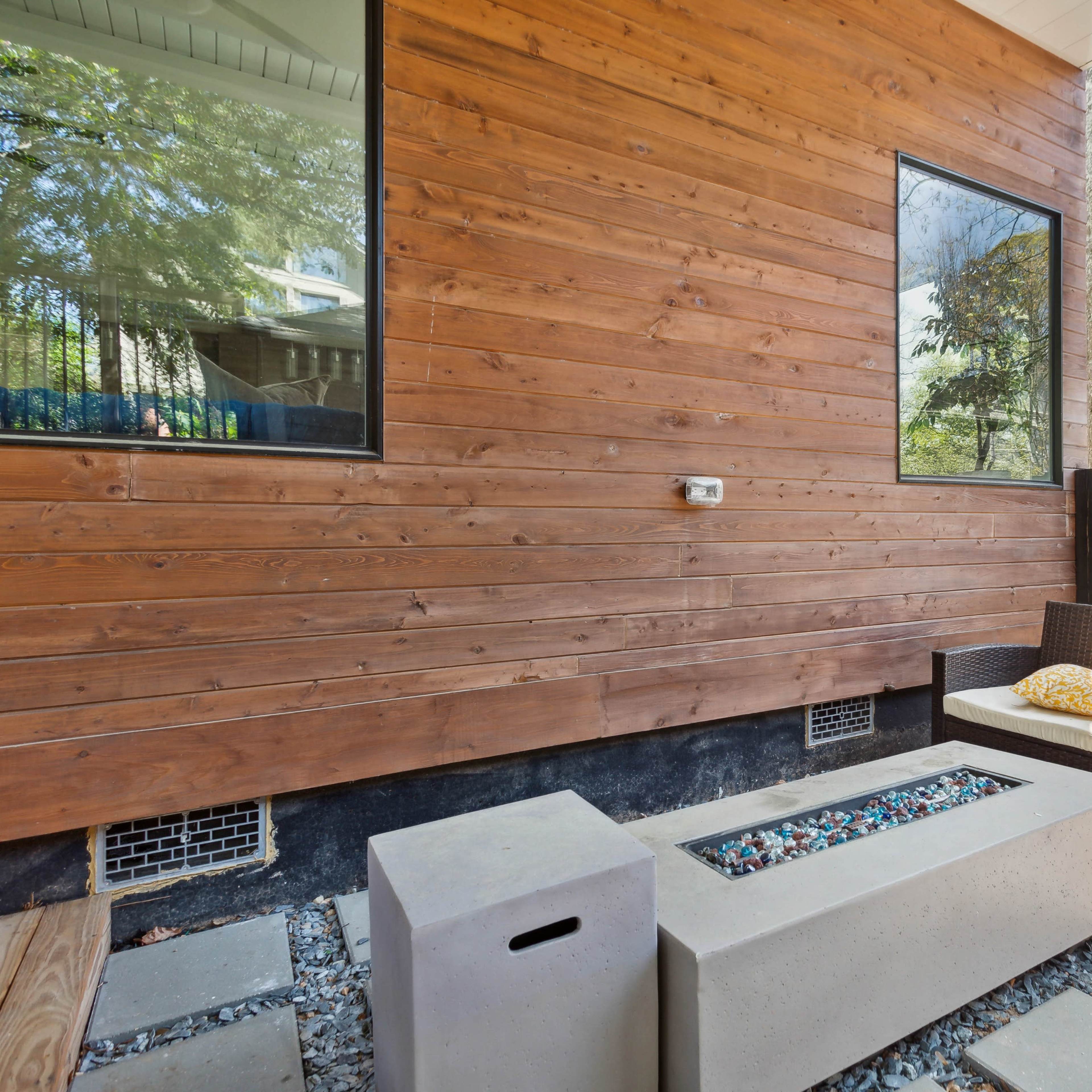 A seating area with a wicker chair and patterned cushions is situated next to a concrete fire pit, set against a wooden wall and surrounded by stone pavers.