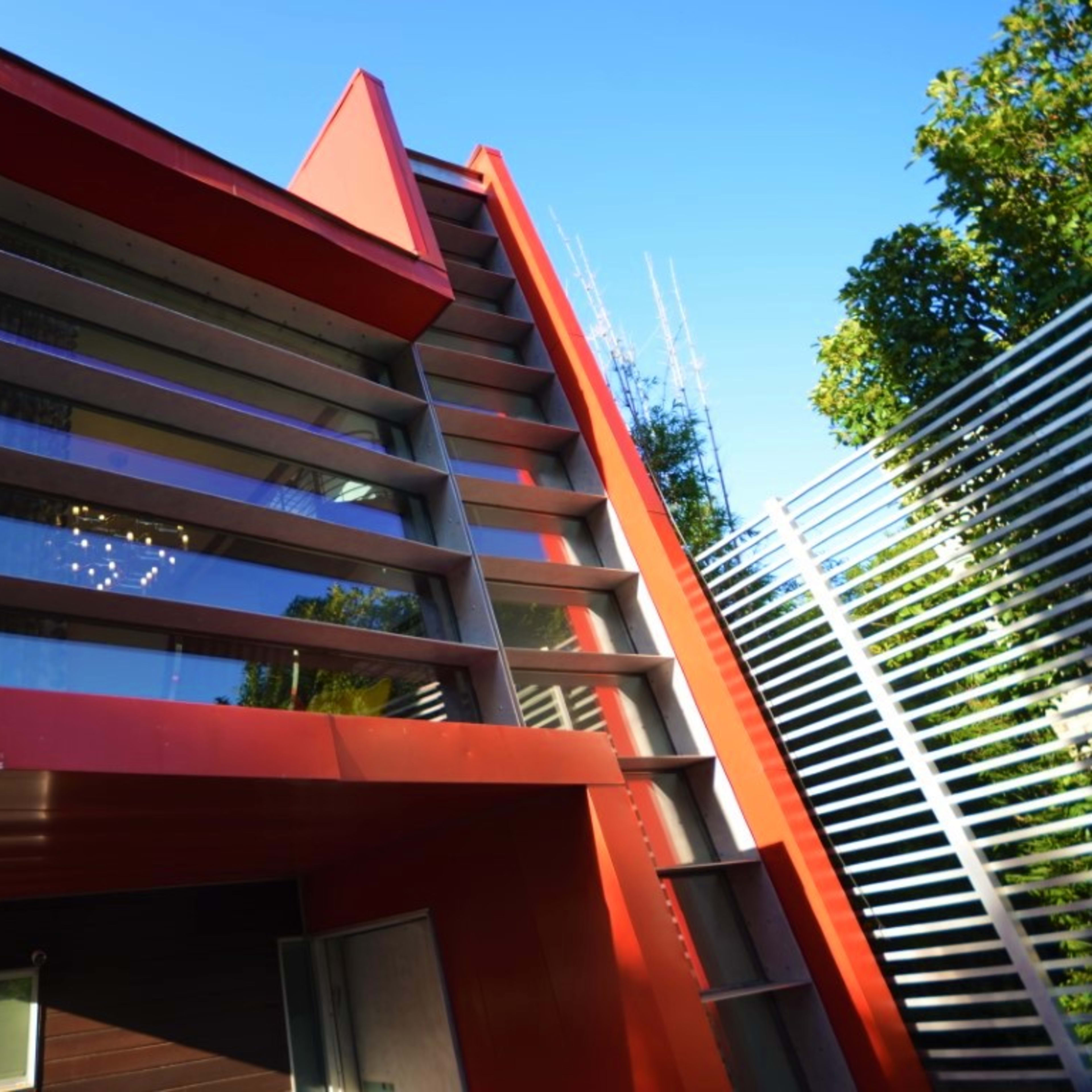A modern red house with large windows and geometric lines, surrounded by trees and a metal fence.