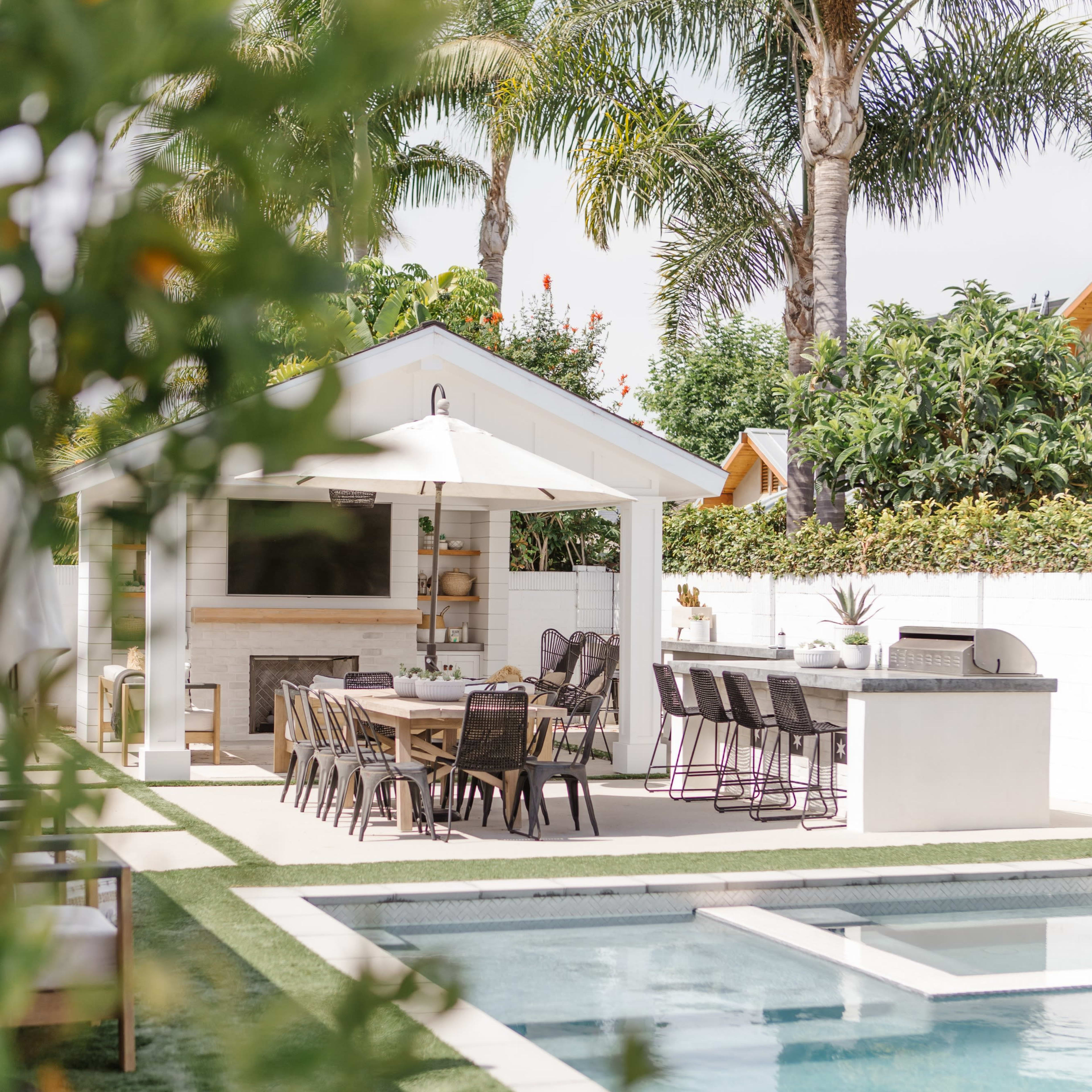 A poolside area features a dining set and a barbecue station surrounded by greenery and palm trees.