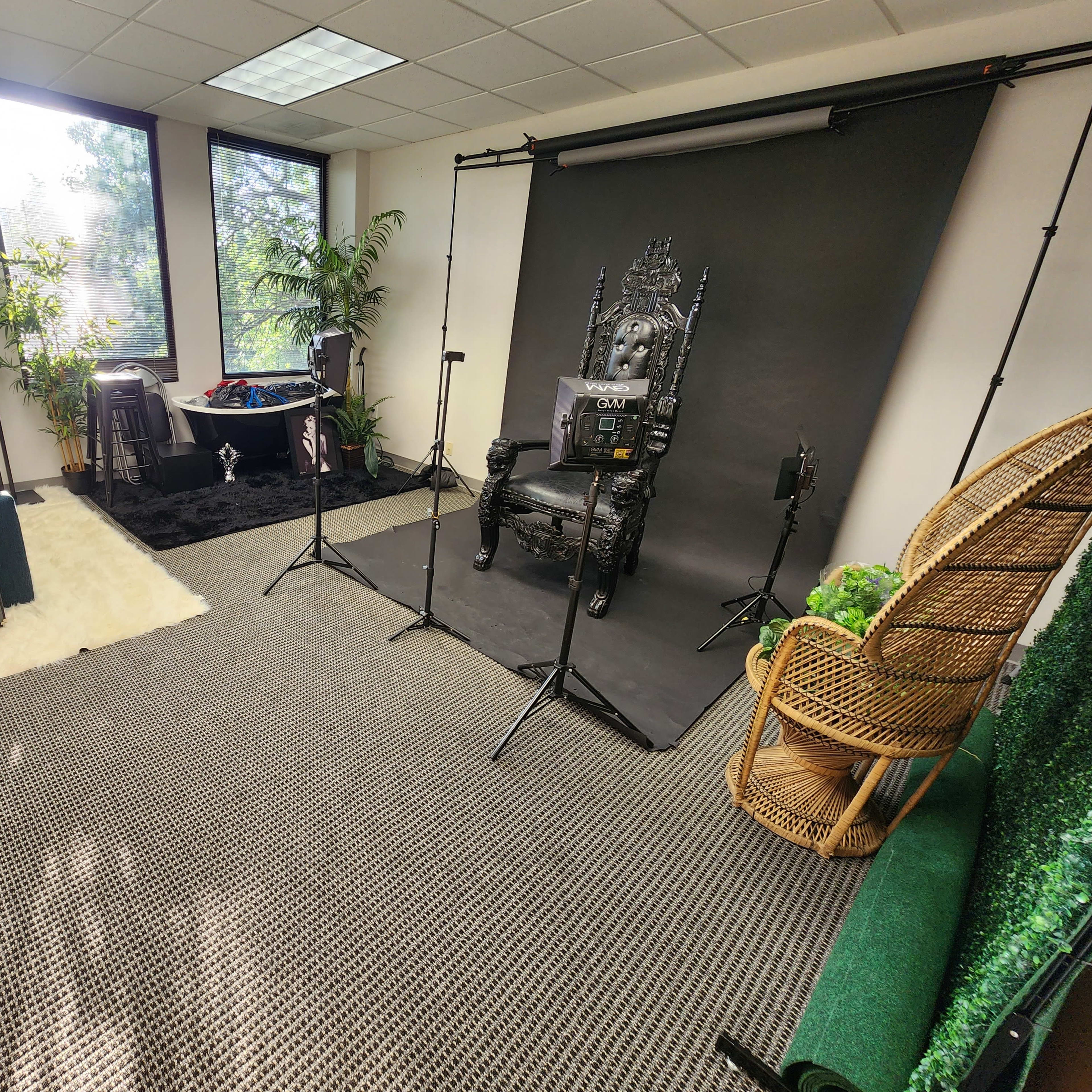 A spacious office studio with a black backdrop set up for photography, featuring a throne-like chair, lighting equipment, and a green plant corner.