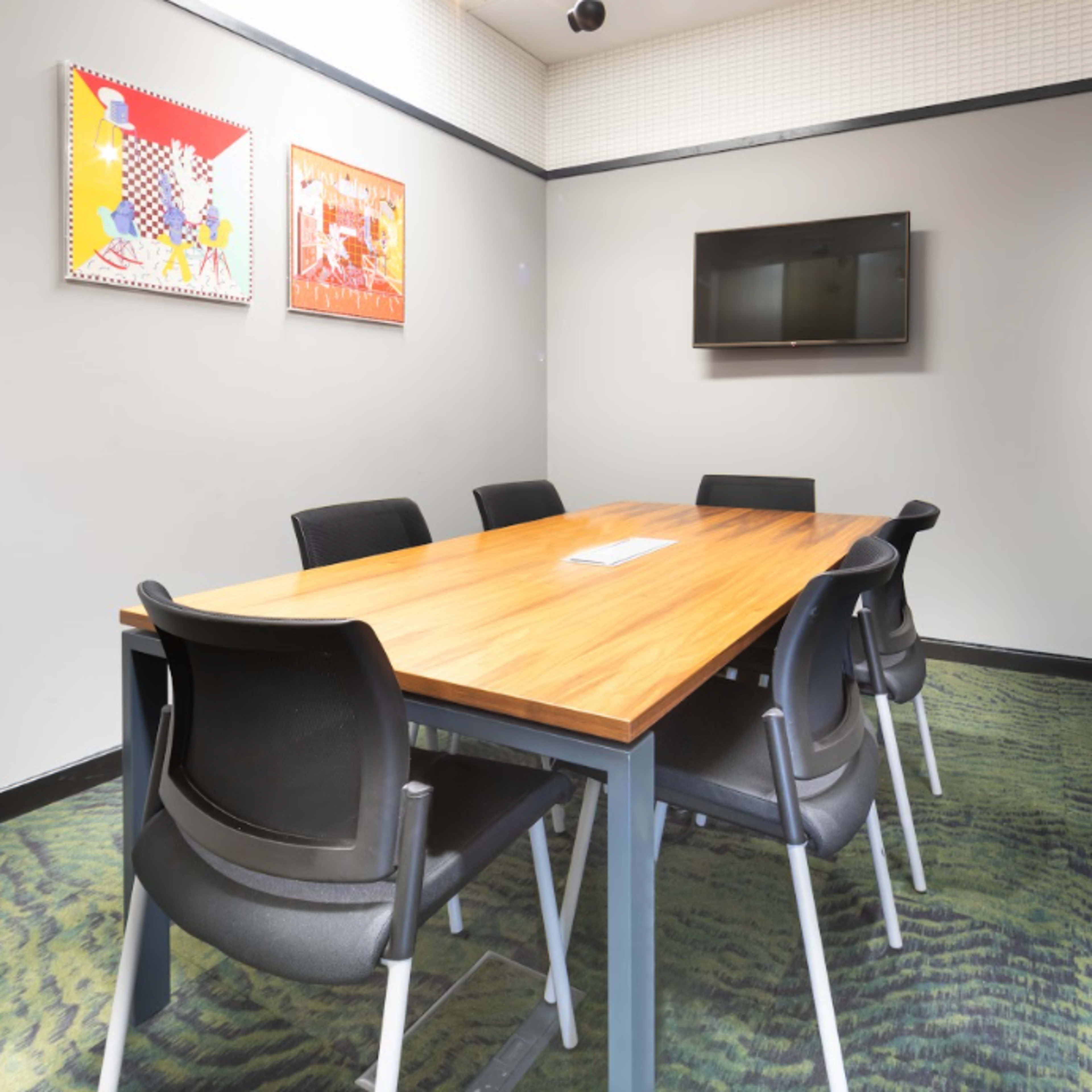 A conference room features a wooden table surrounded by black chairs, with two framed artworks on the wall and a flat-screen TV mounted opposite the table.