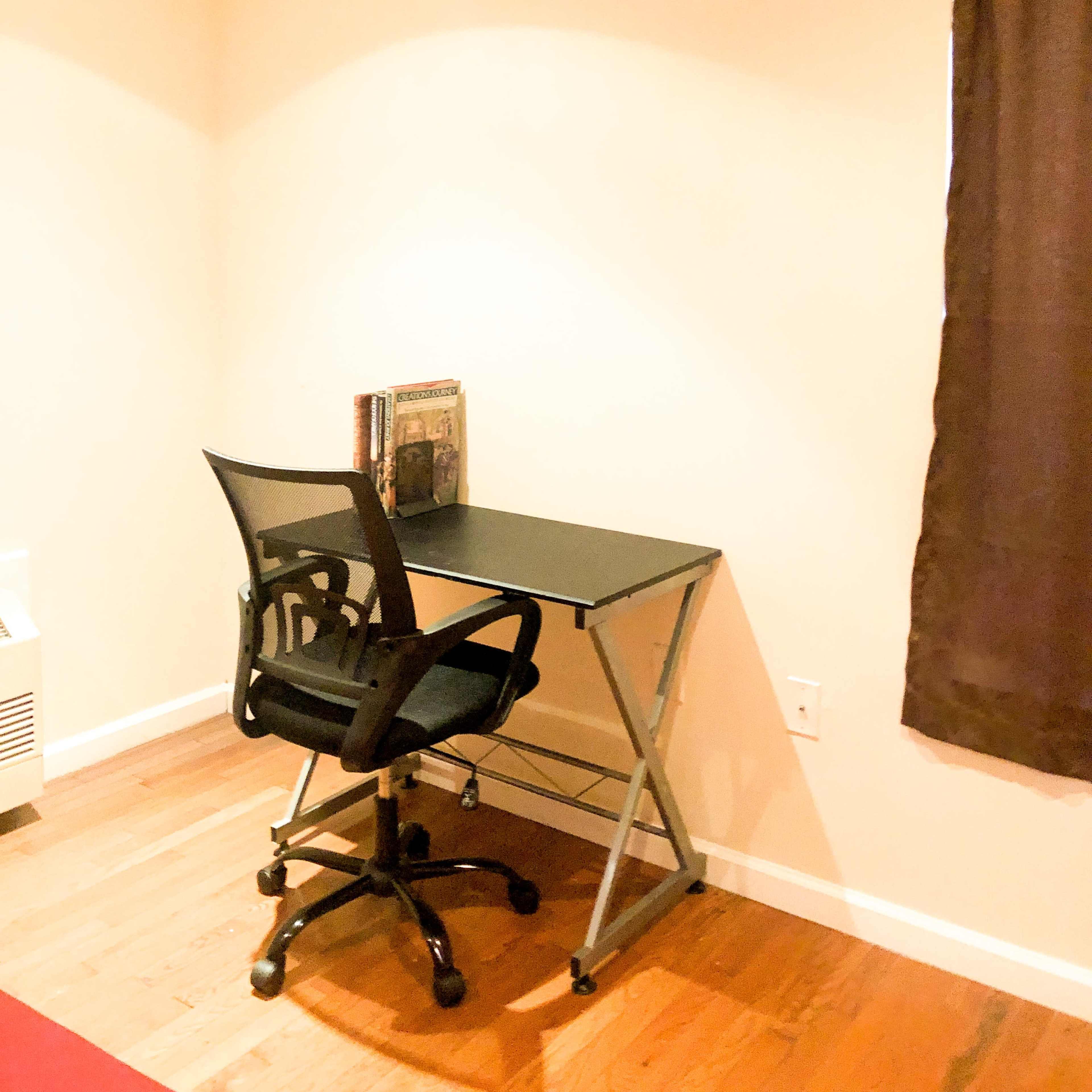 A black office chair is positioned in front of a small desk against a light-colored wall, next to a window covered with a brown curtain.