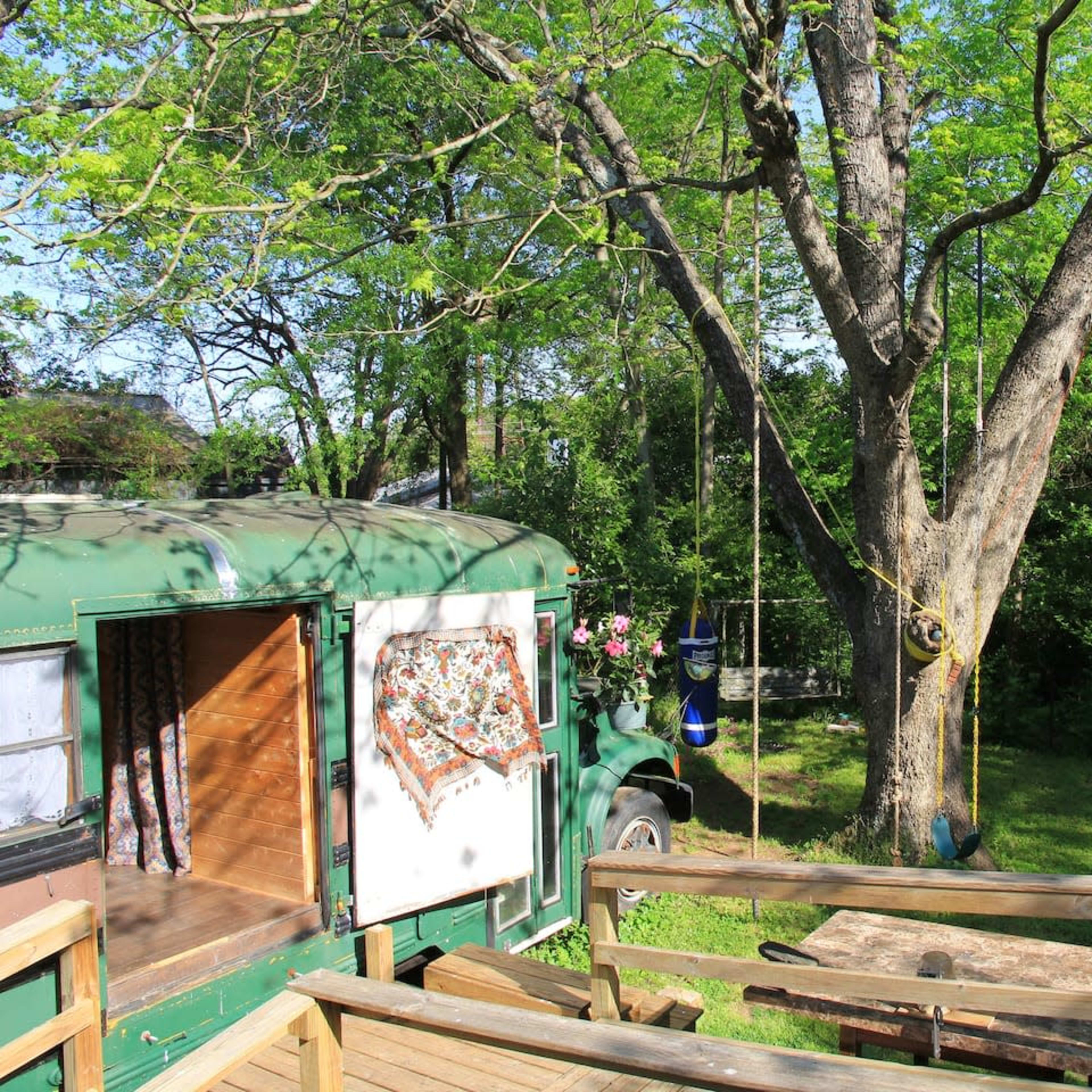 A green bus is parked beside a large tree in a grassy area with a wooden deck and a fire pit nearby.