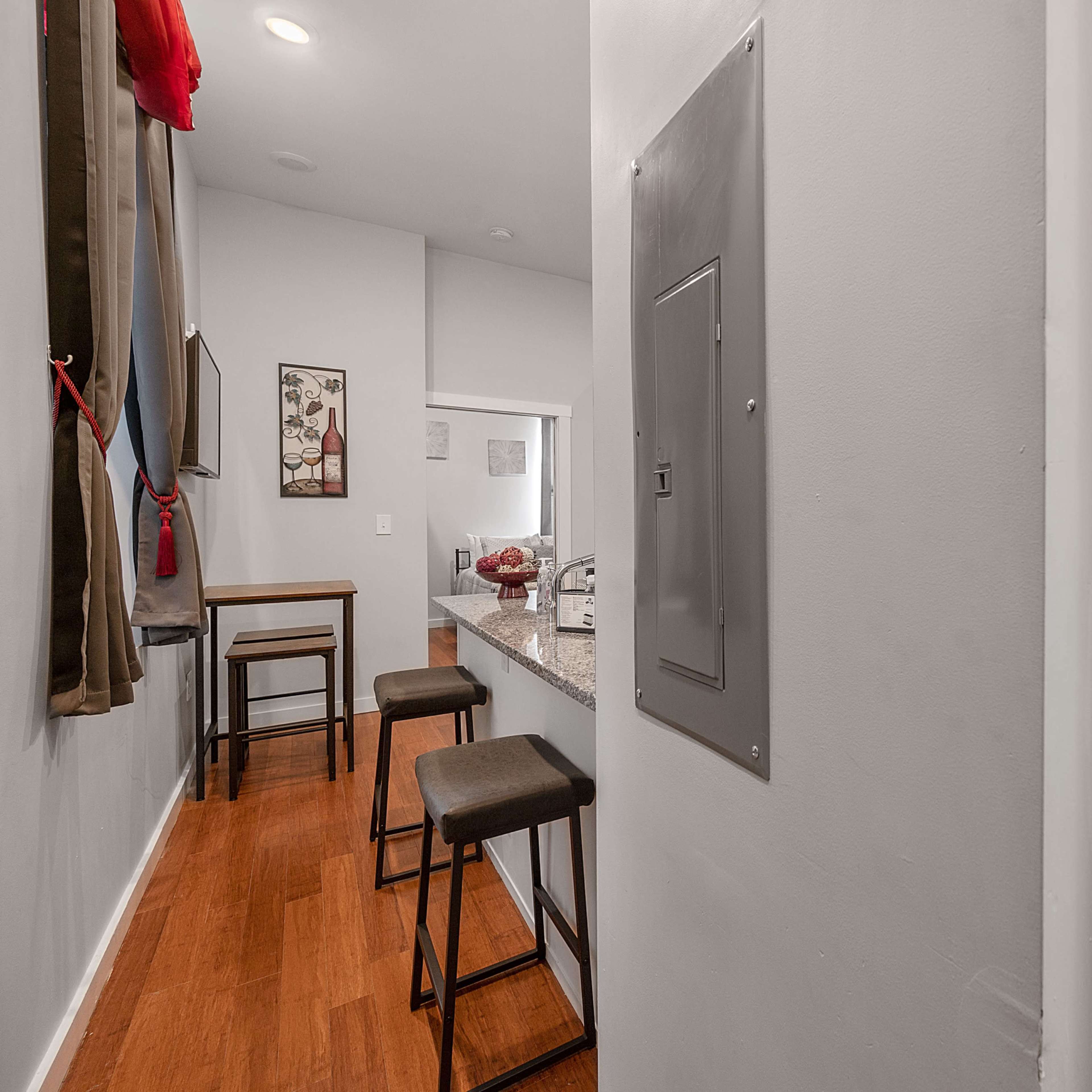A hallway leading to a kitchen area with a countertop and bar stools, a wall-mounted electrical panel, and a bathroom door with a shower curtain visible.