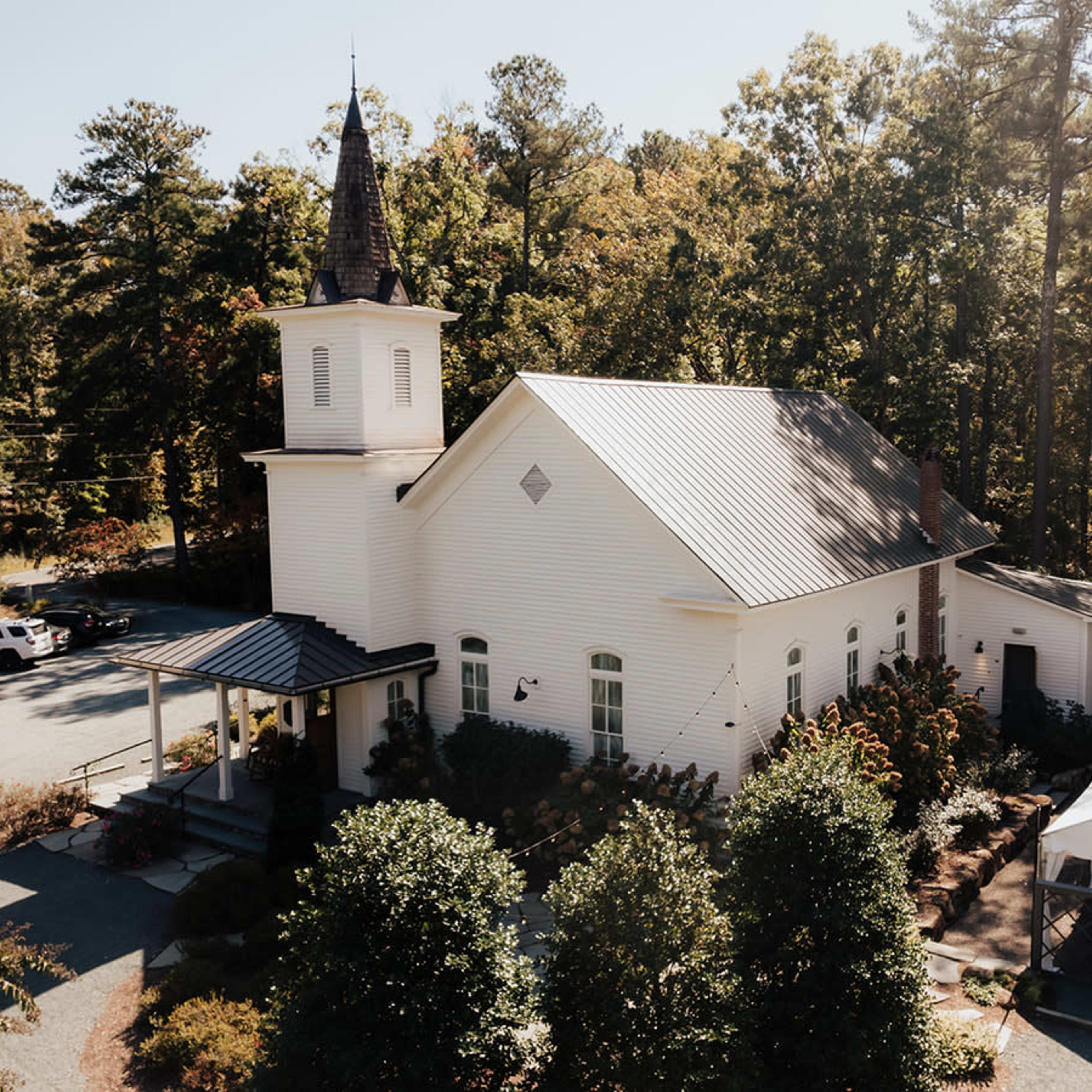 A white church with a steeple is nestled among trees, adjacent to a large tent and a gravel parking area.