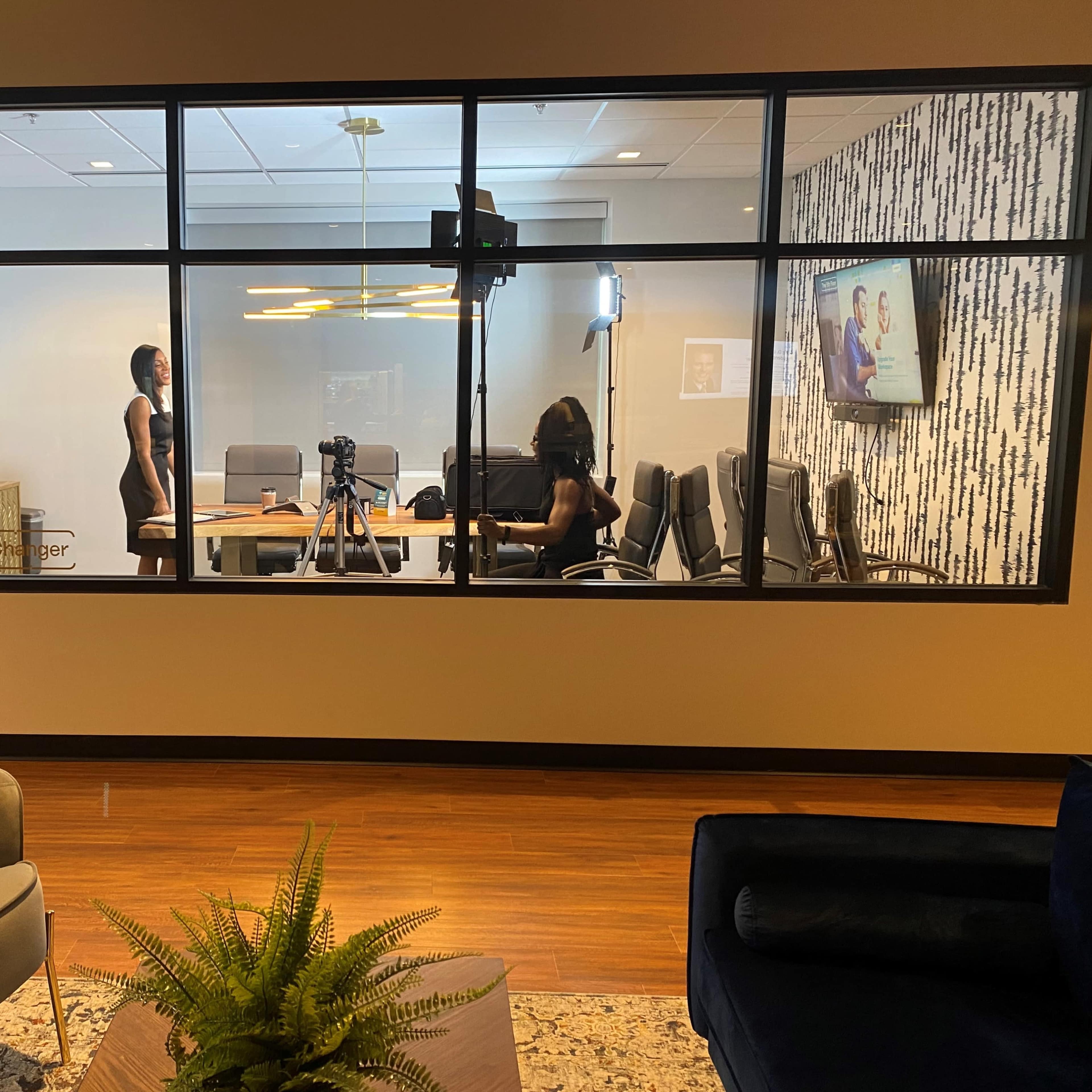 A modern meeting room with glass walls features a table surrounded by chairs, a camera setup, and a person standing while another person sits in front of a screen.