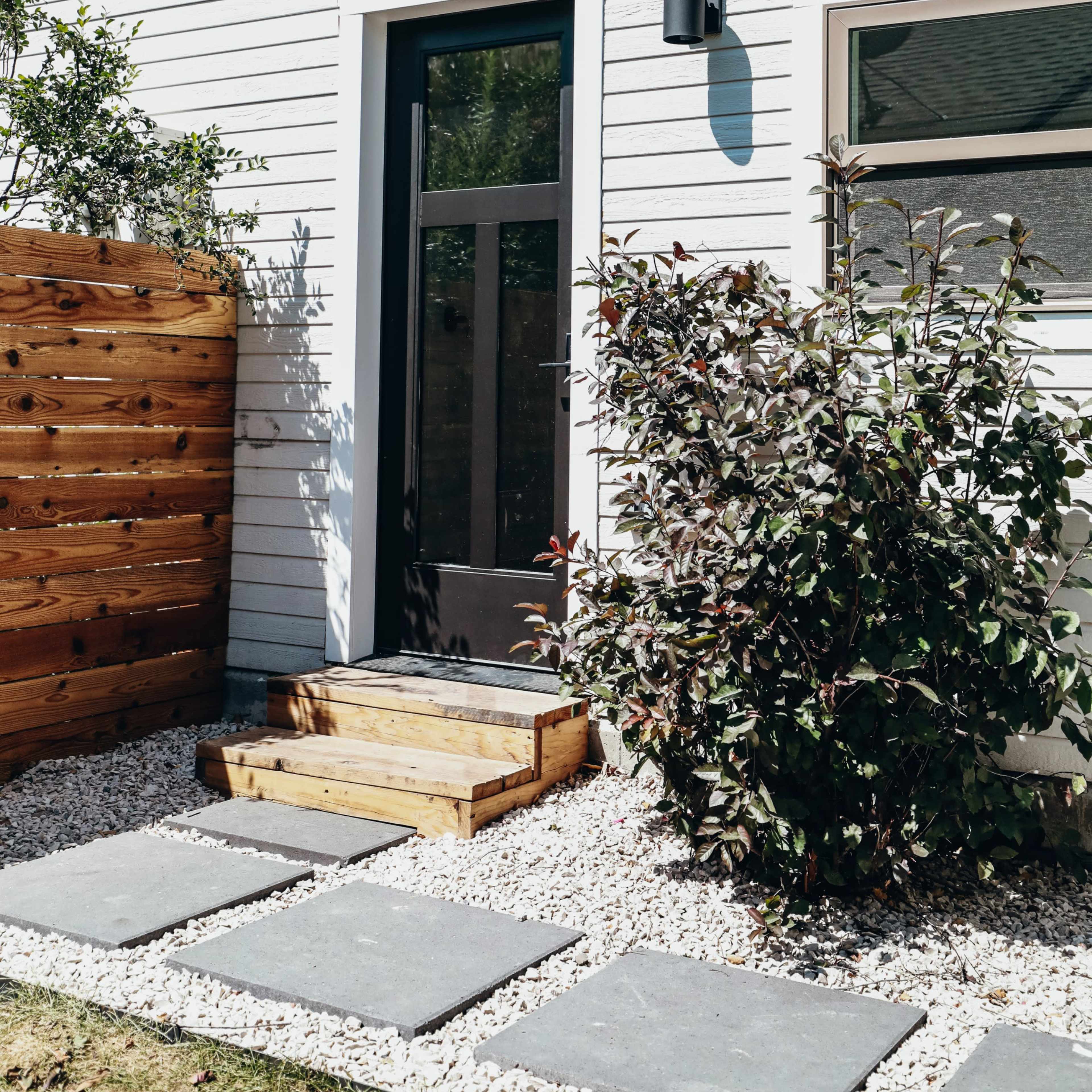 A wooden step leads to a door framed by a bush and flanked by a wooden fence, set against a textured white wall.