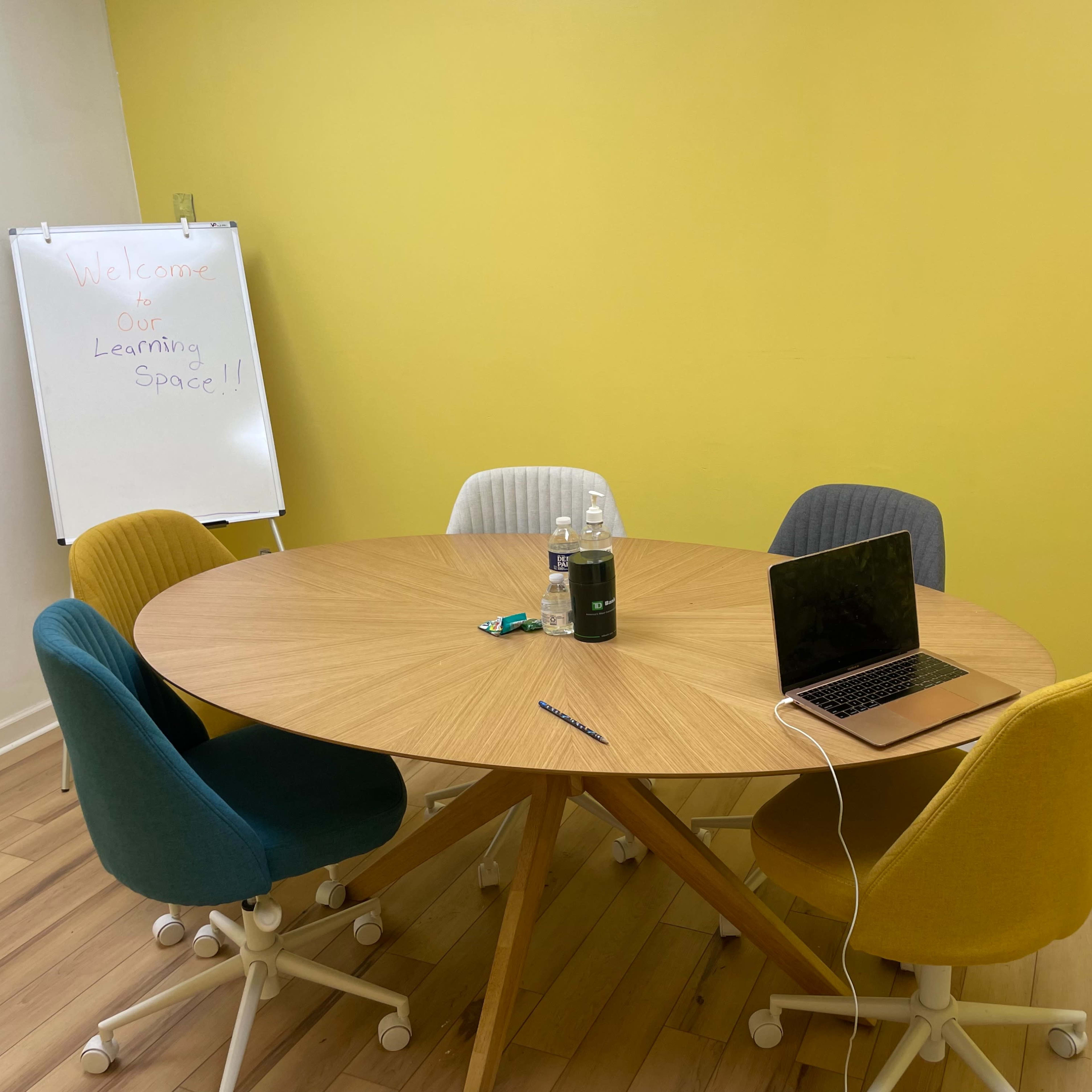 A round wooden table surrounded by five chairs in a room with a yellow wall and a whiteboard.