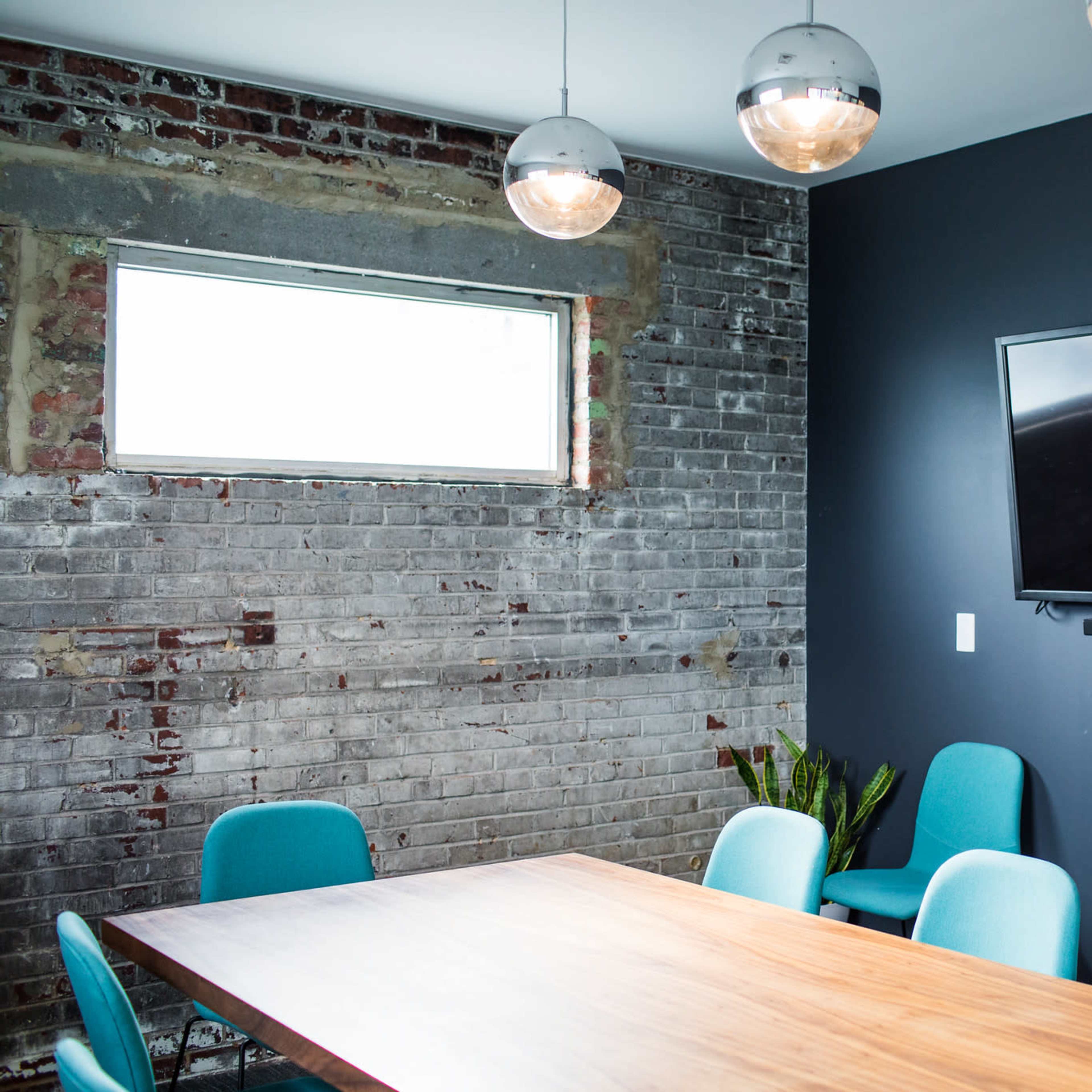 A conference room features a wooden table surrounded by blue chairs, with a flat-screen TV mounted on a dark wall and a window providing natural light.
