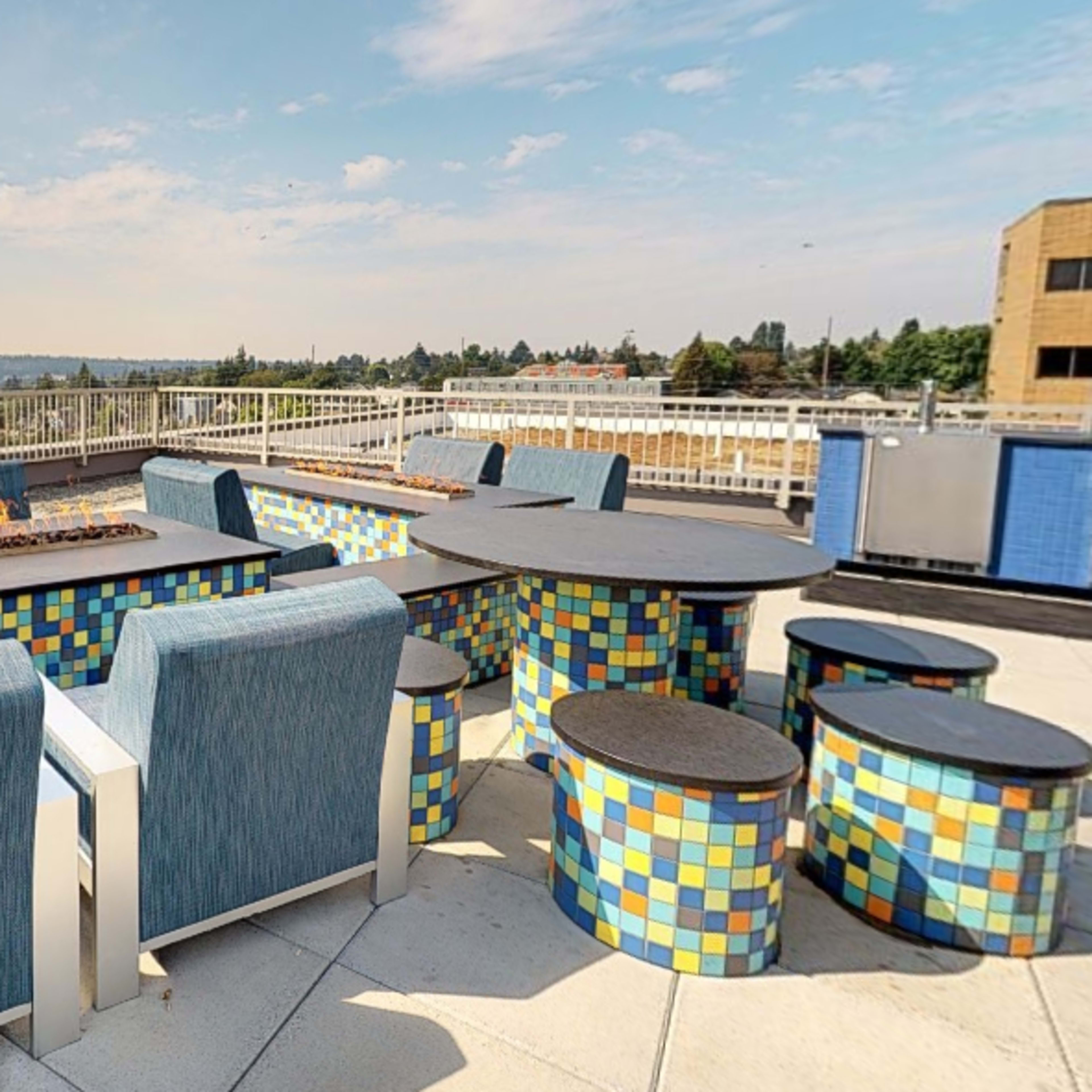 A rooftop terrace with a fire pit surrounded by colorful tiled seating and a panoramic view of a city skyline.
