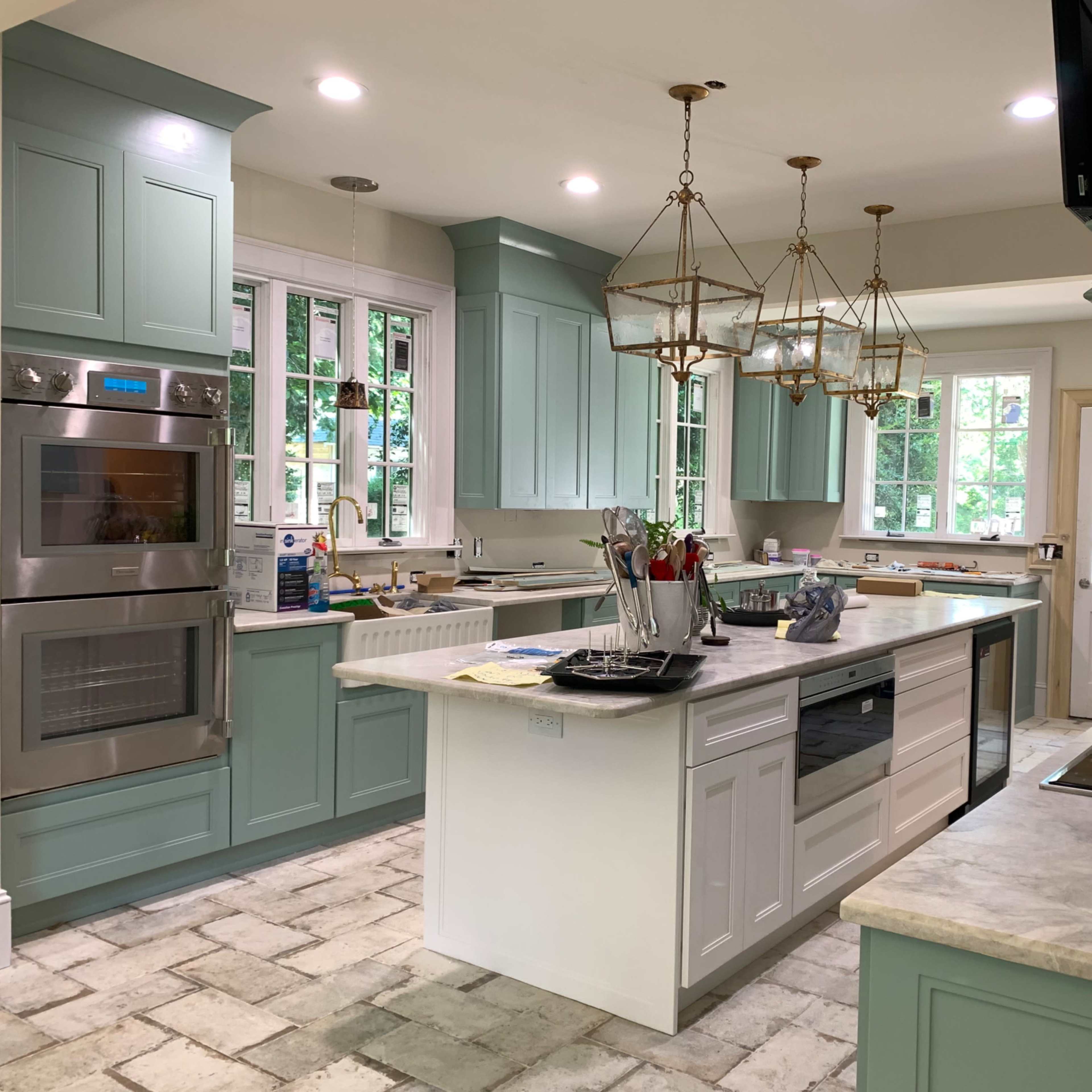 A modern kitchen featuring light green cabinetry, a central island with a countertop, and multiple light fixtures hanging above.