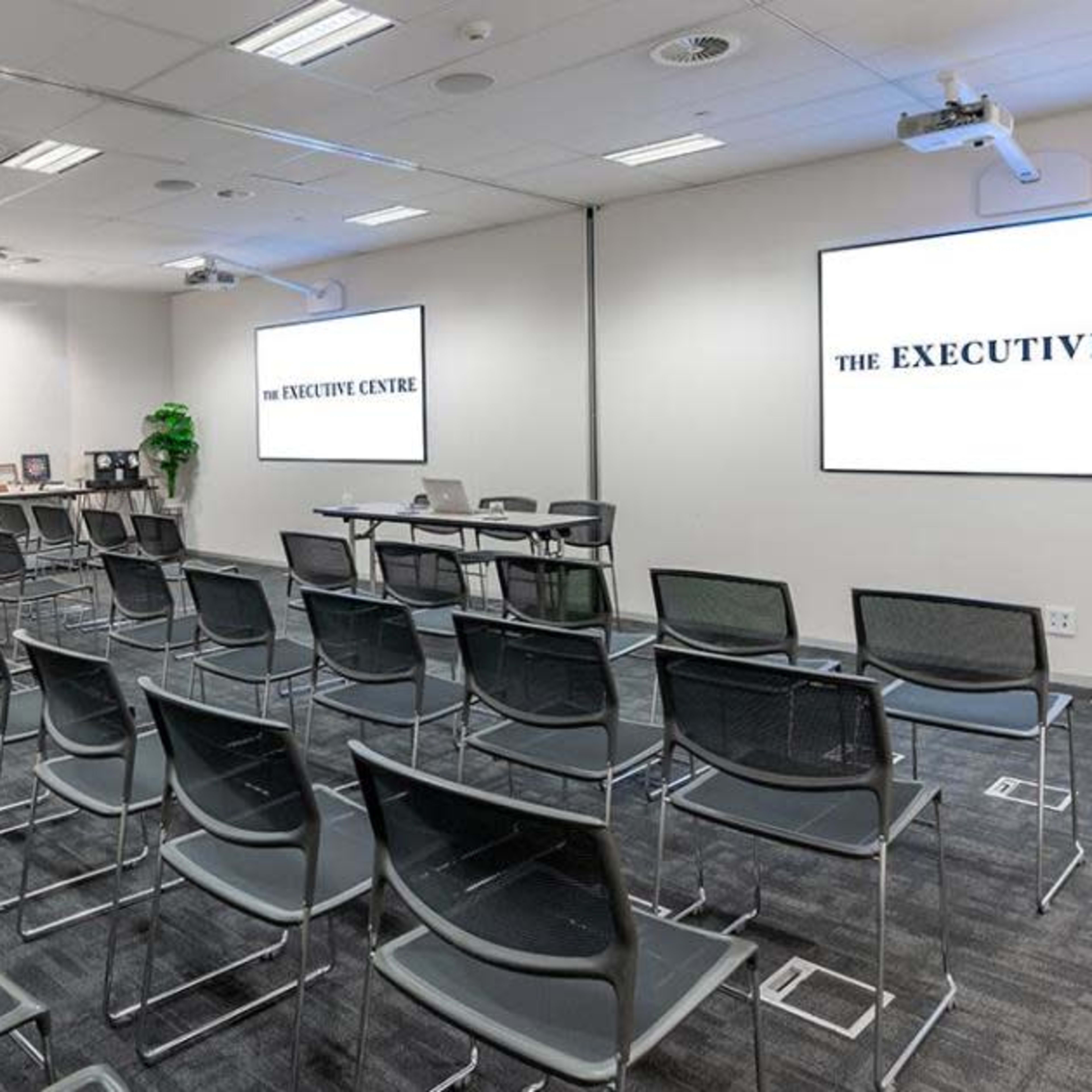 A modern conference room with rows of black chairs, a projection screen displaying "THE EXECUTIVE CENTRE," and a refreshment table along one wall.