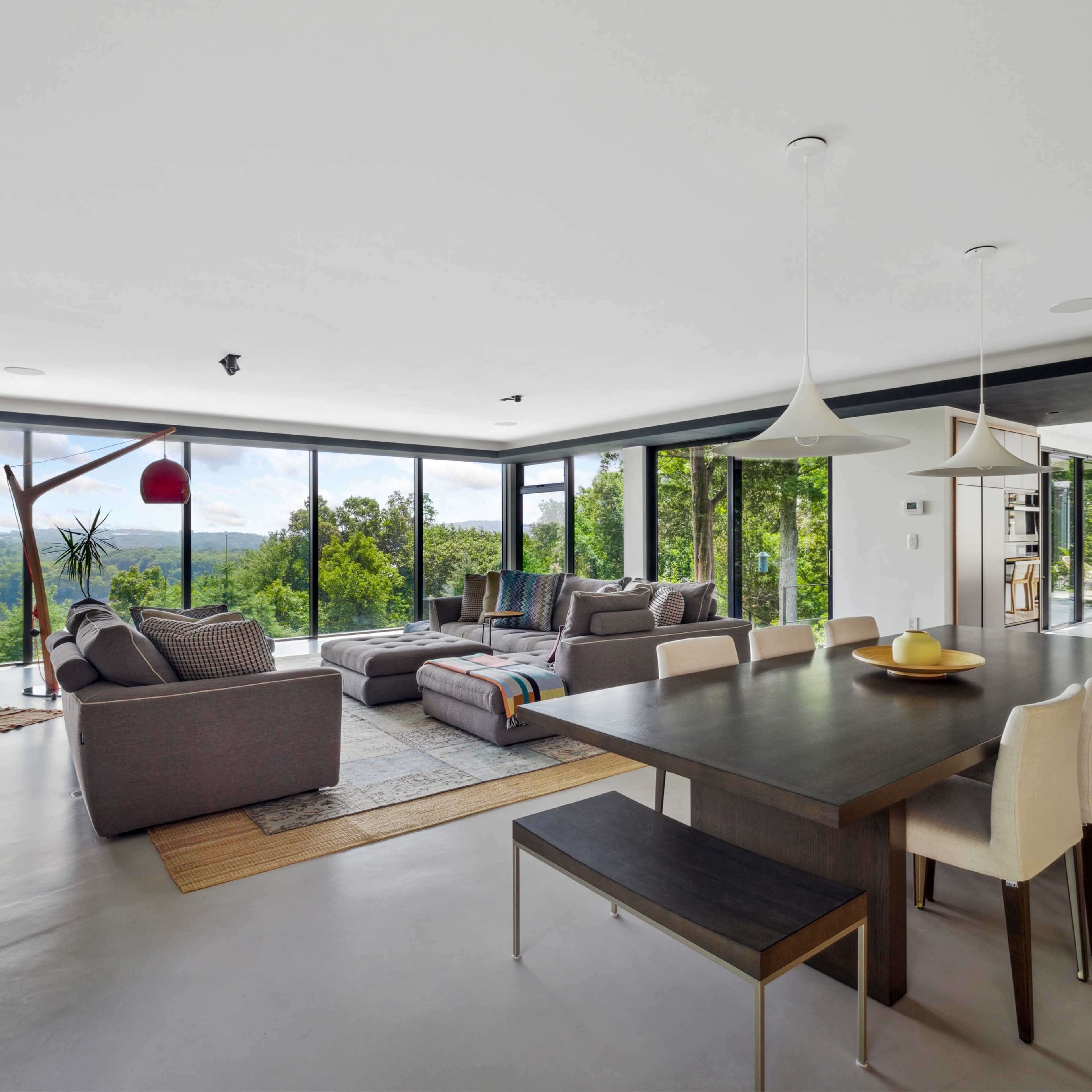 A modern living space with a seating area featuring large windows overlooking a green landscape and a dining table with chairs.