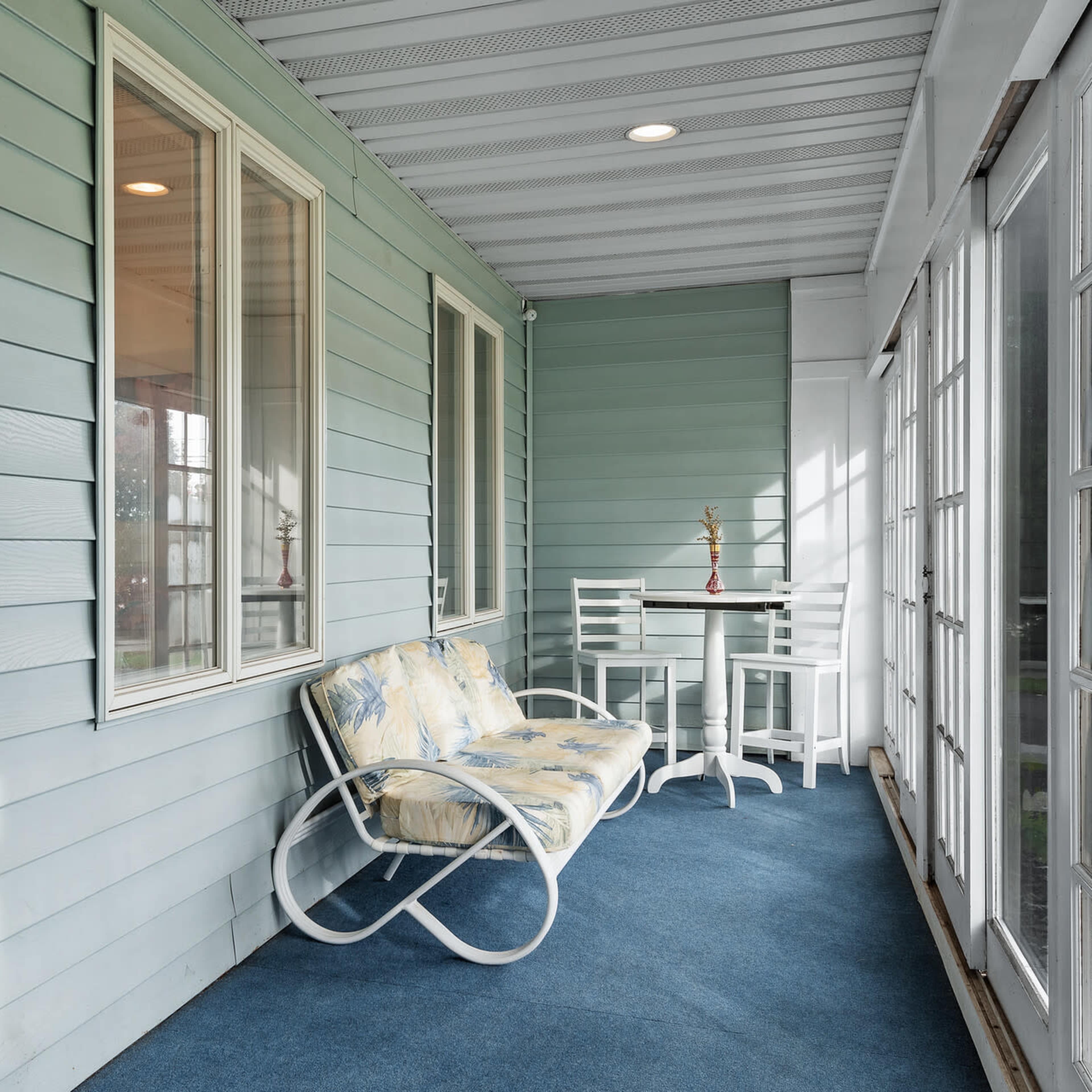 A porch area with a blue wall, two windows, a small table, and a white sofa with a floral cushion.