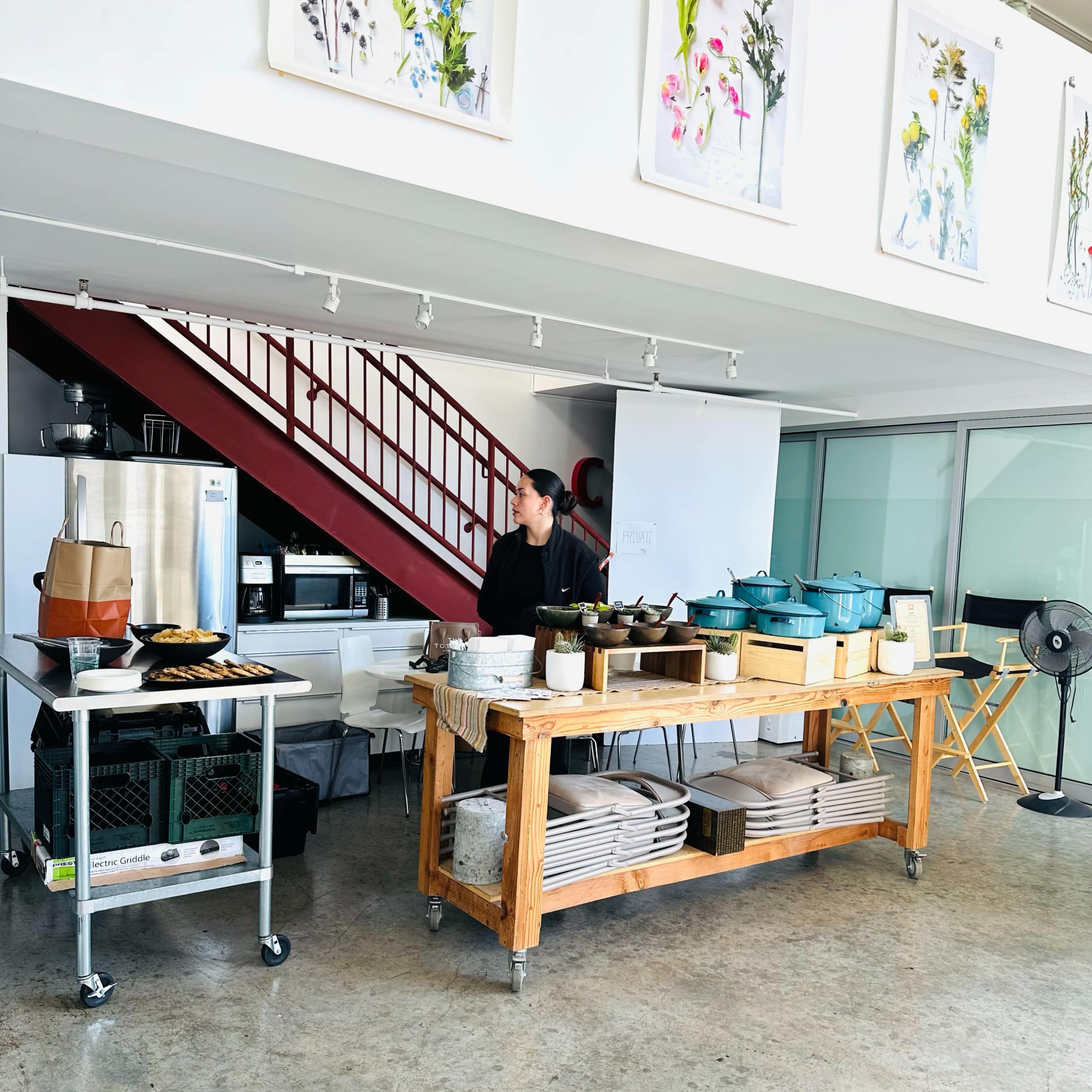 A woman stands in a modern kitchen space with a wooden work table and various cooking utensils, under framed artworks of flowers.