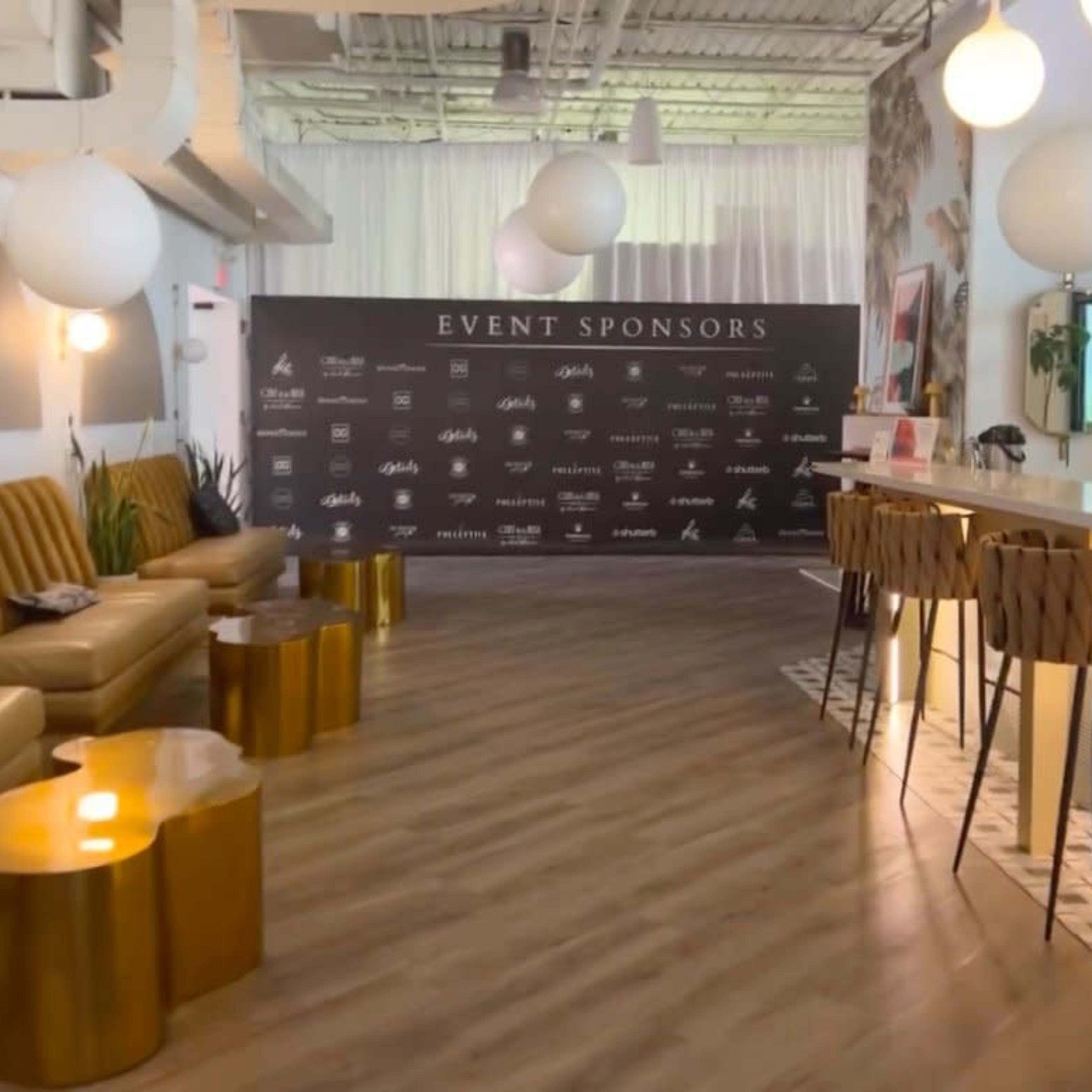 A modern event space with a long brownie leather sofa, round gold accent tables, a bar area, and a backdrop displaying event sponsors.